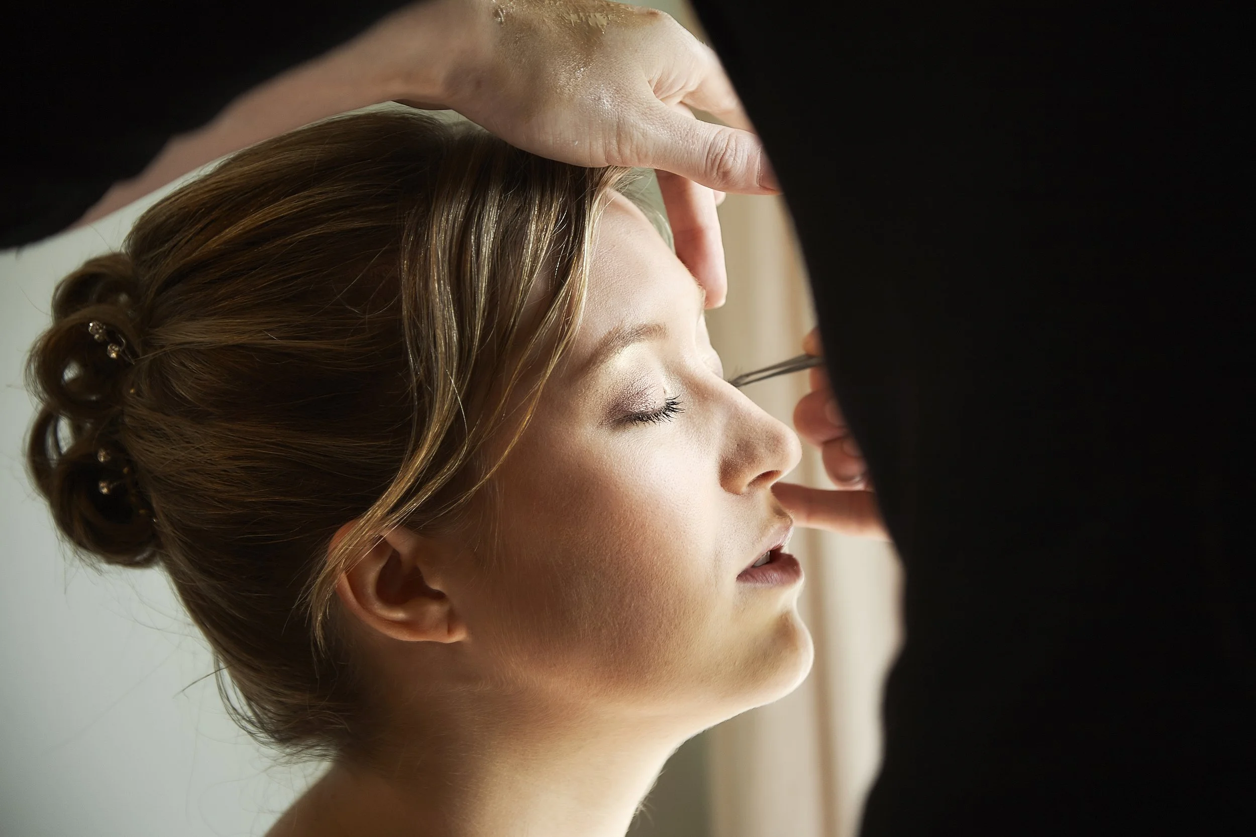 A makeup artist applying makeup to a woman's face with her eyes closed, indoors with a blurred background.