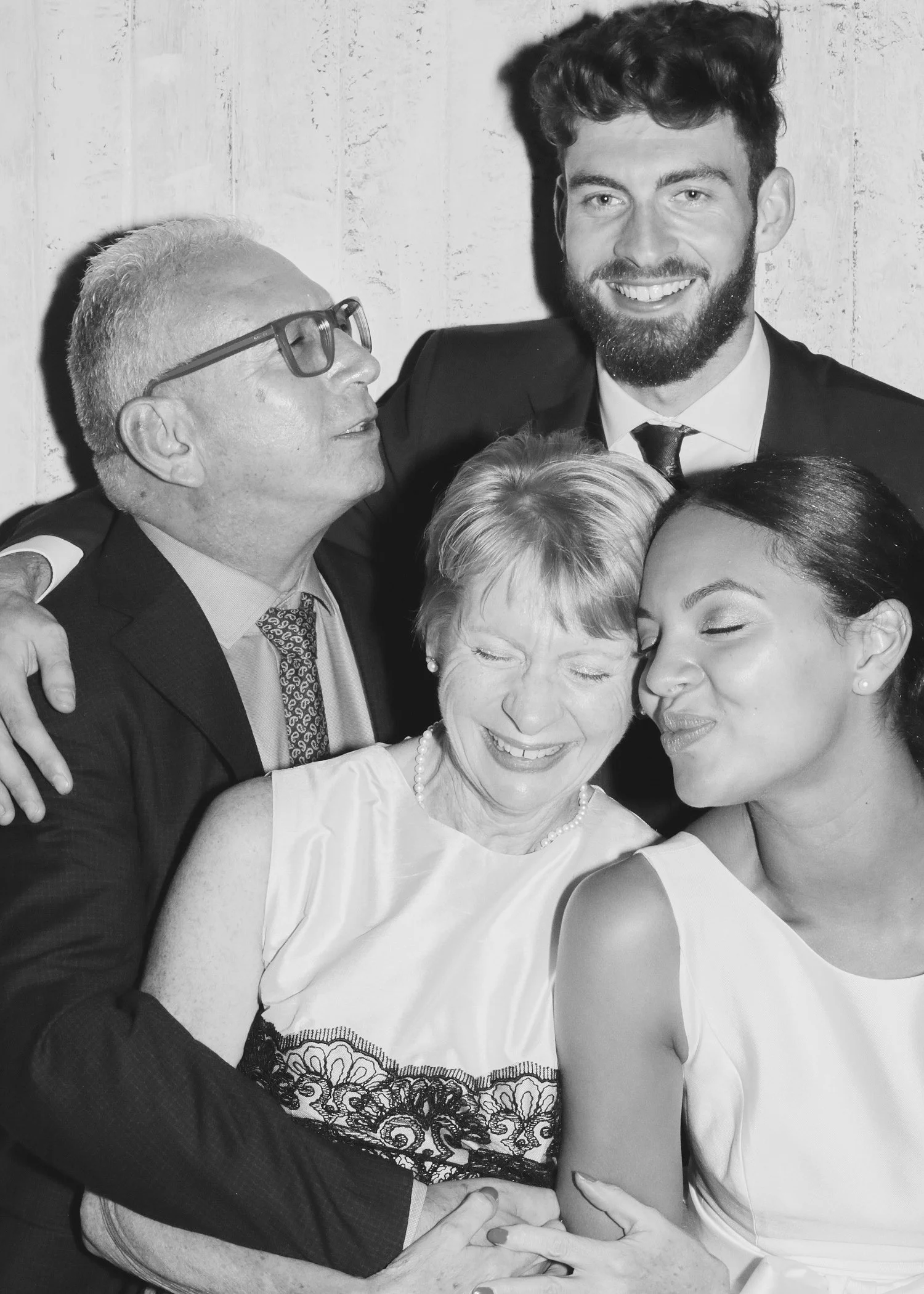 A black and white photo of five people, hugging and smiling. An elderly woman with earrings and a pearl necklace is in the center, being hugged by a woman in a sleeveless dress, a young man in a suit, and a man with glasses and gray hair.