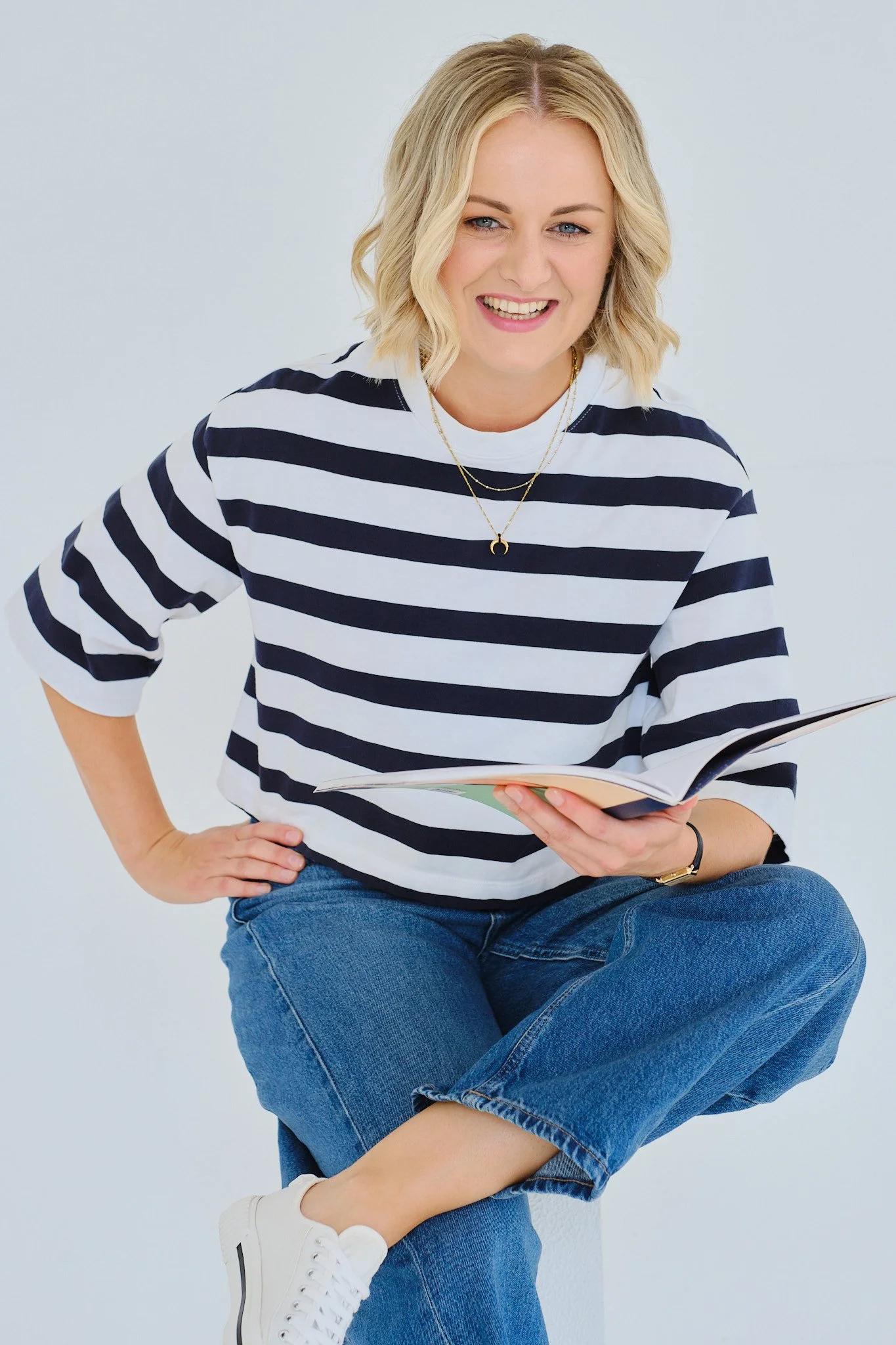 A woman with blonde hair, wearing a black and white striped shirt, blue jeans, and white sneakers, is sitting against a white background, smiling and holding a book.
