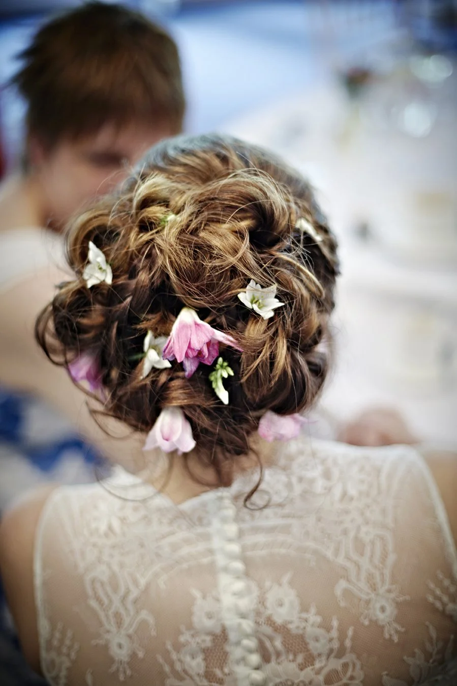 A woman with curly, braided hairstyle adorned with white, pink, and green flowers, seen from behind, wearing a lace dress, at a wedding or formal event.