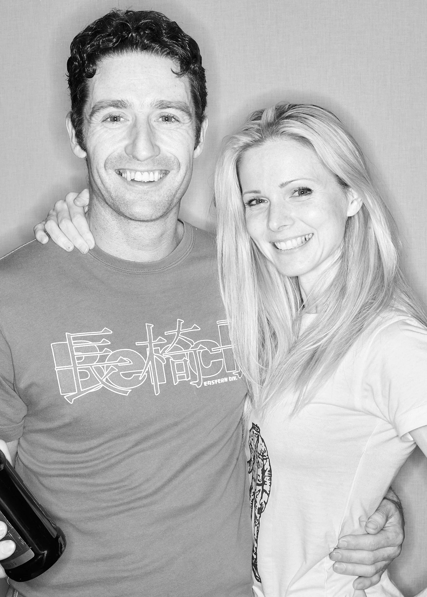 A black and white photo of a smiling man and woman embracing each other, with the woman's arm around the man's shoulder, and the man holding a smartphone in his hand.