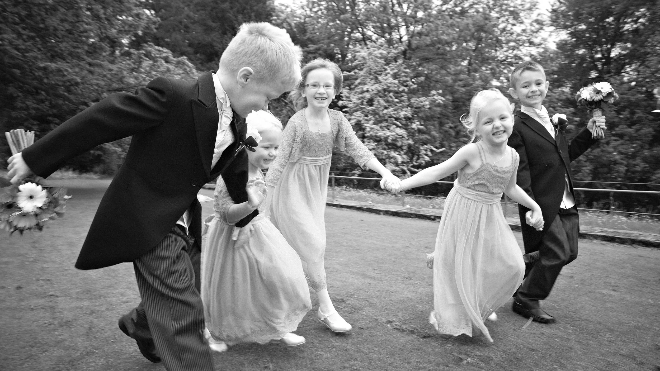 Black and white photo of children and a young man holding hands and running outdoors, smiling and enjoying themselves at a park or garden setting.