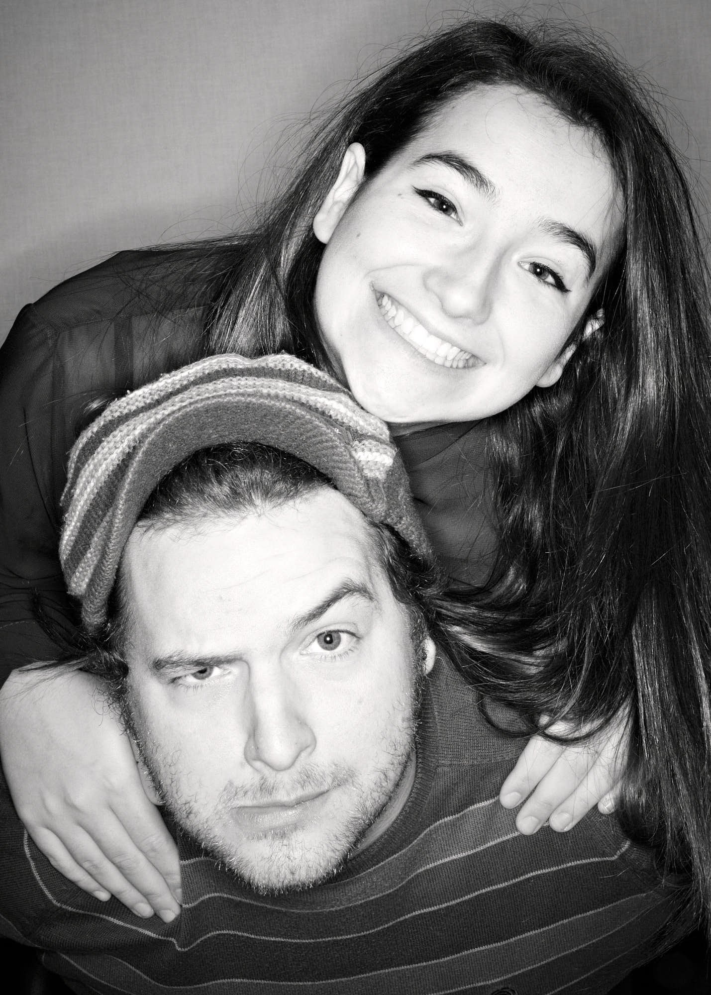 A smiling woman with long dark hair hugging a young man with short dark hair and light skin, both in close-up black and white portrait.