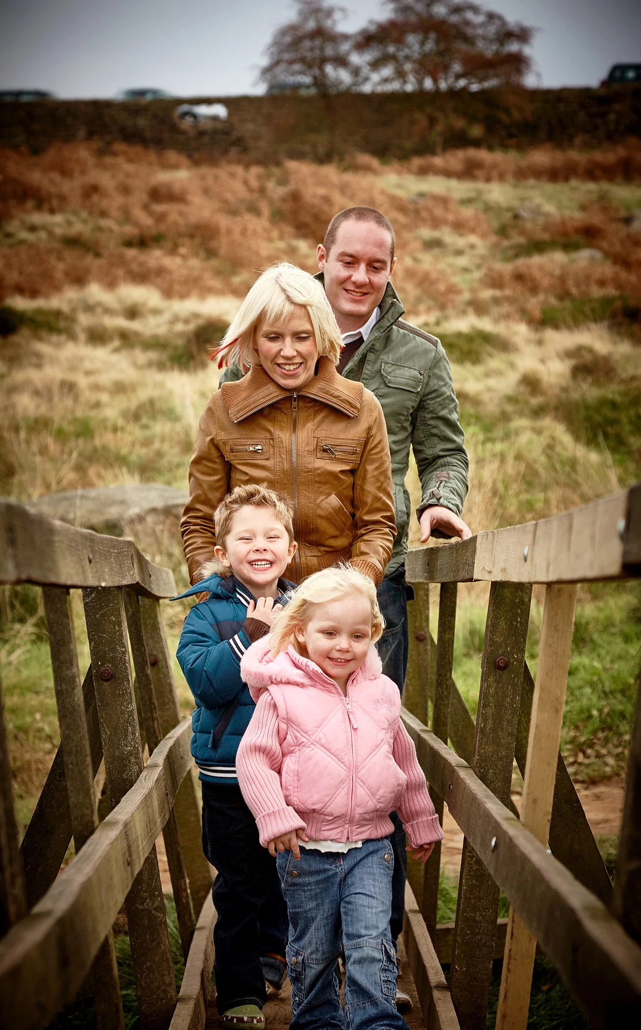 A group of children and adults walking across a wooden footbridge in a rural outdoor setting with brown and green grass and a tree in the background.