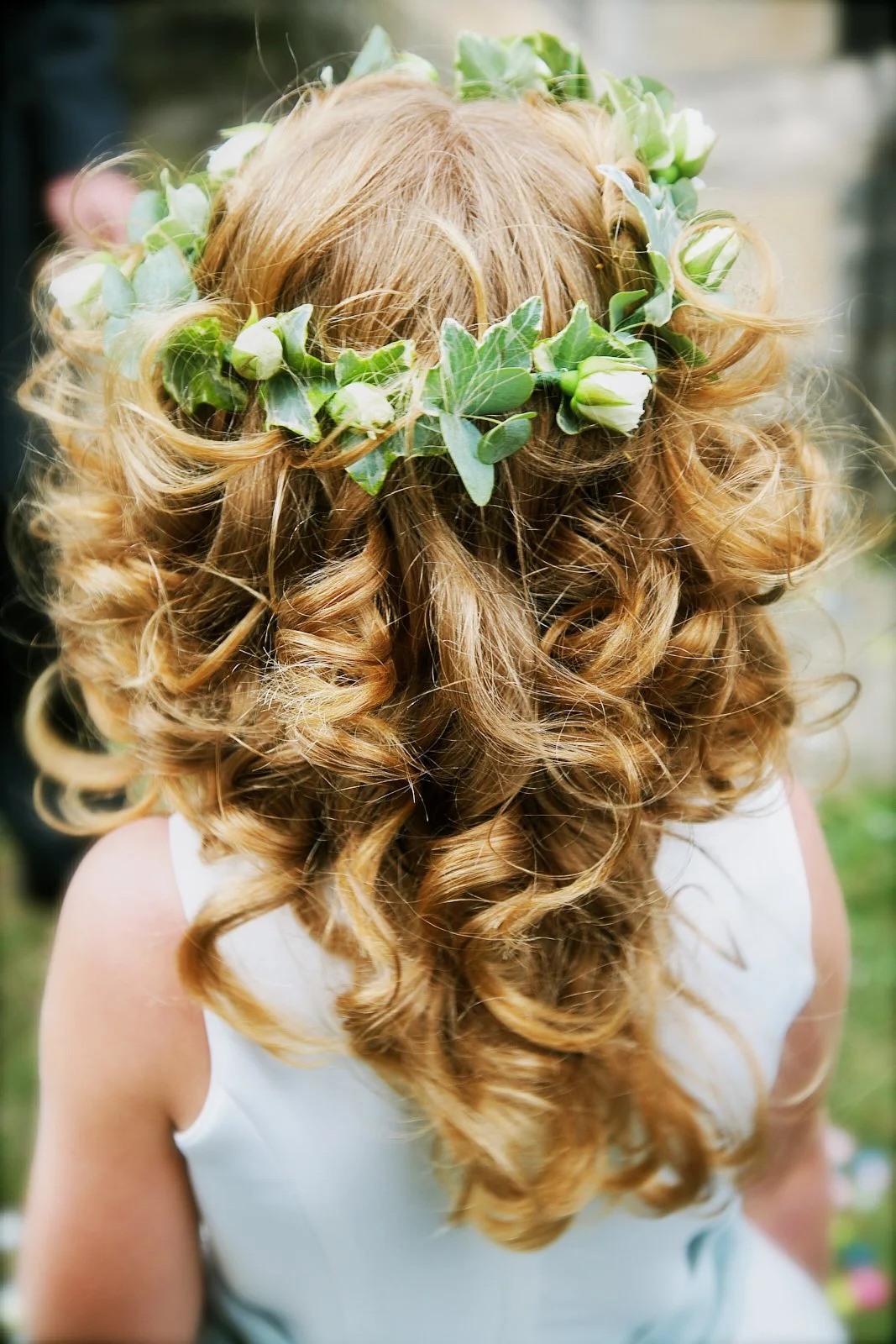 Back view of a woman with curly red hair wearing a floral crown made of green leaves and white flowers, dressed in a white sleeveless dress, outdoors.