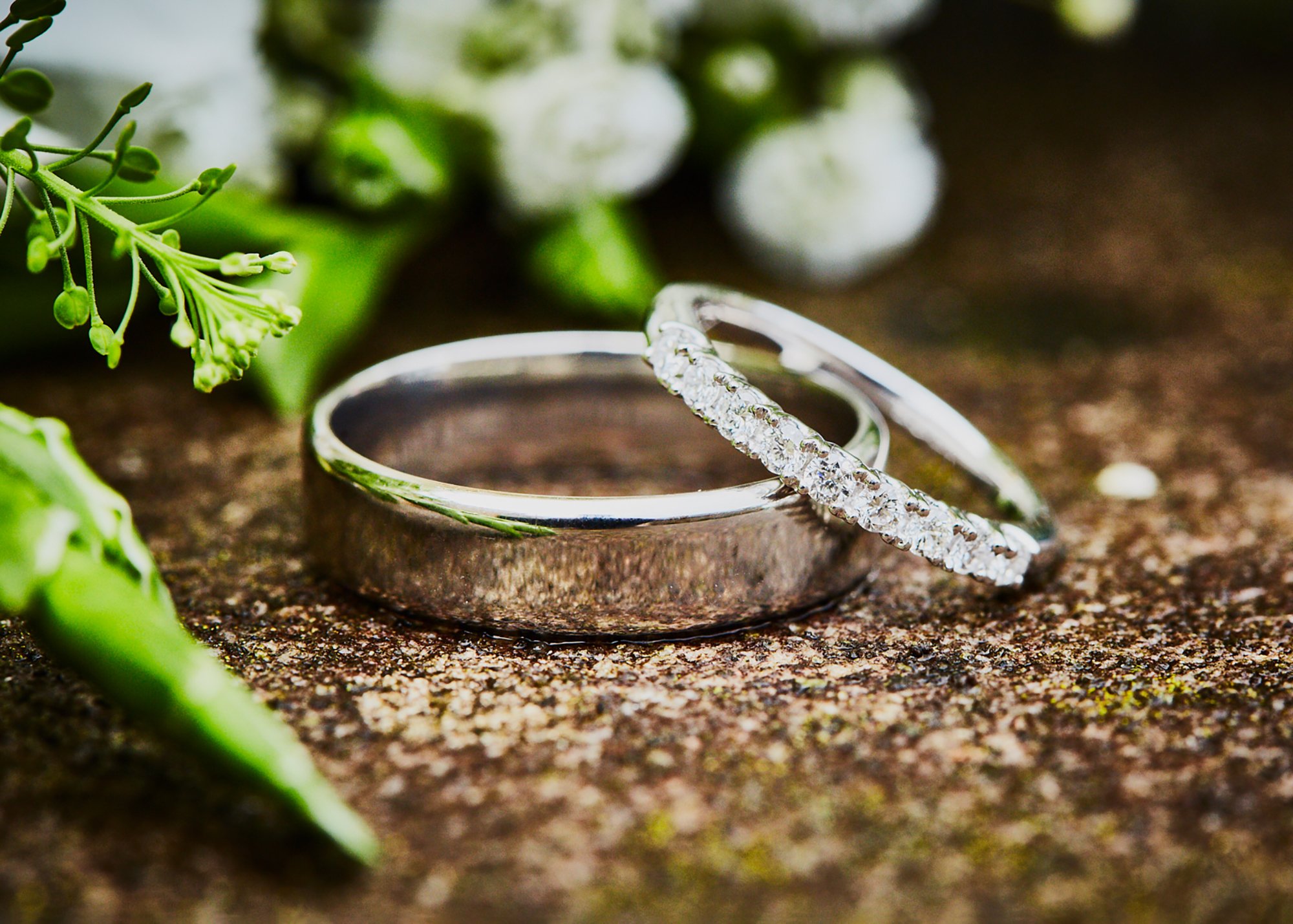 Close-up image of a silver ring with a large central diamond and smaller diamonds on the band, along with a matching ring, placed on a rustic wooden surface with green leaves and small white flowers in the background.
