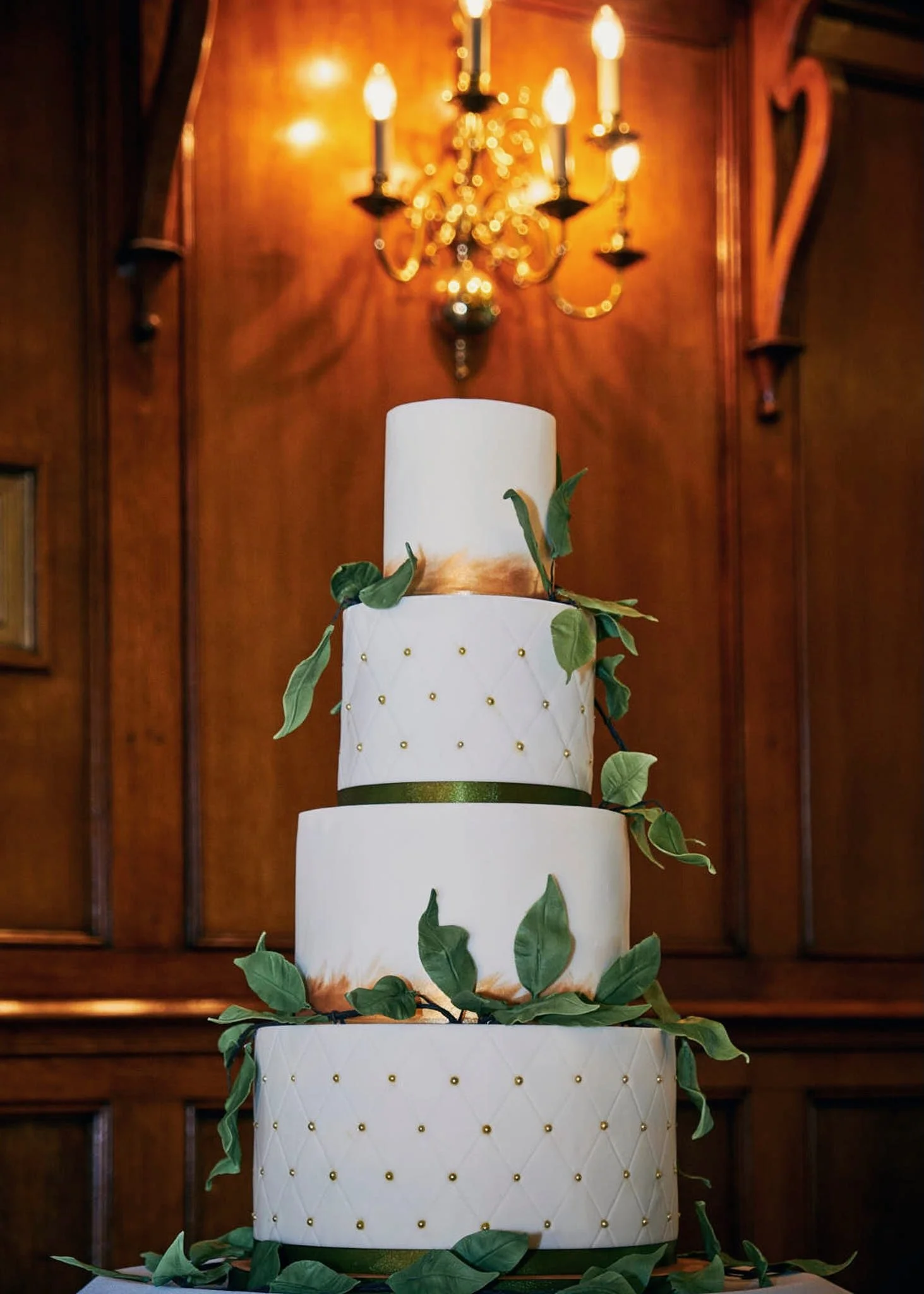 A multi-tiered white wedding cake decorated with green leaves and gold accents, set against a wooden background with a chandelier
