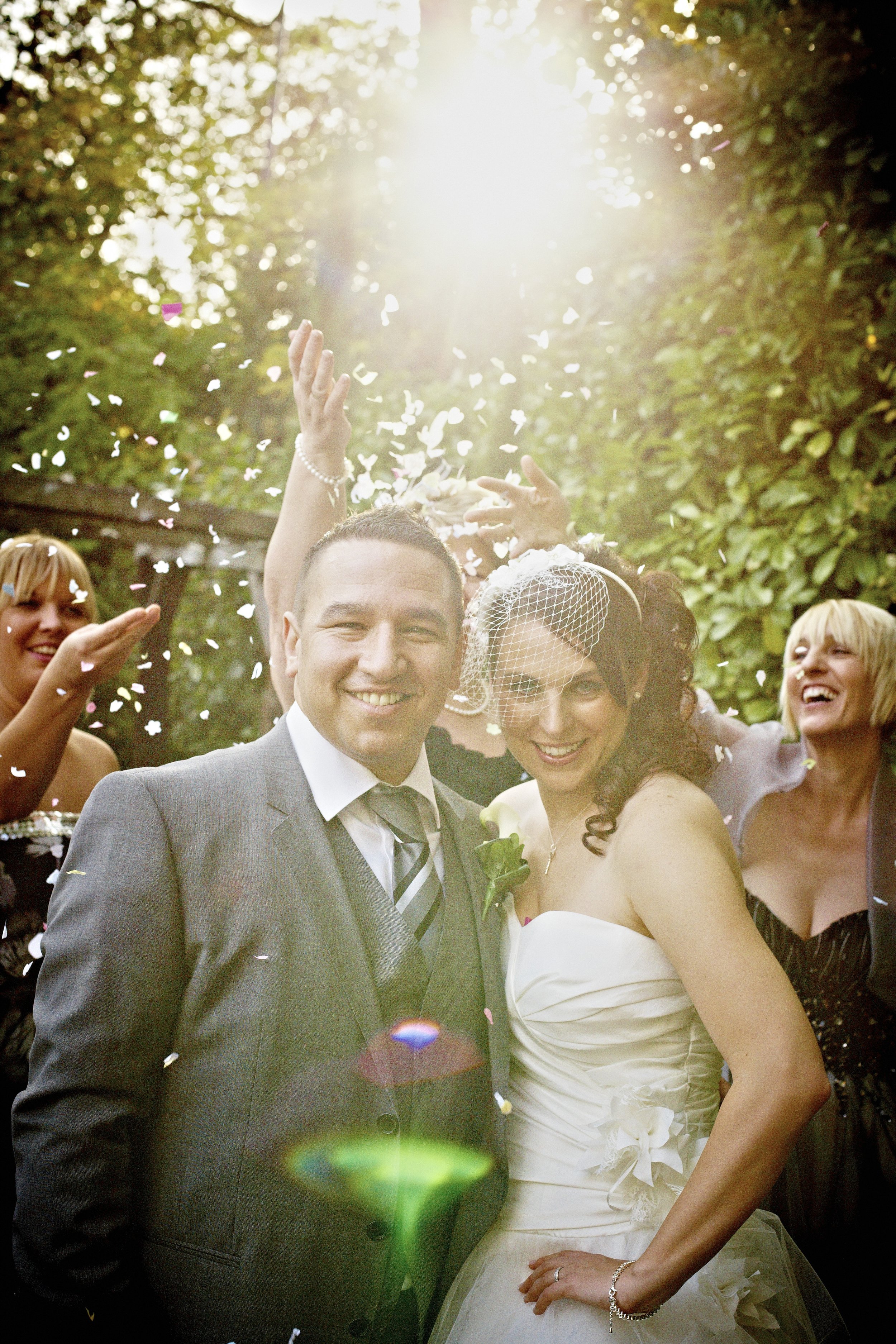 A group of people celebrating at a wedding, with a bride and groom in the center, surrounded by friends throwing confetti, in outdoor setting with sunlight filtering through trees.