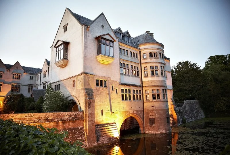 A historic castle-like building illuminated at dusk, reflecting in a nearby water moat, with trees in the background.