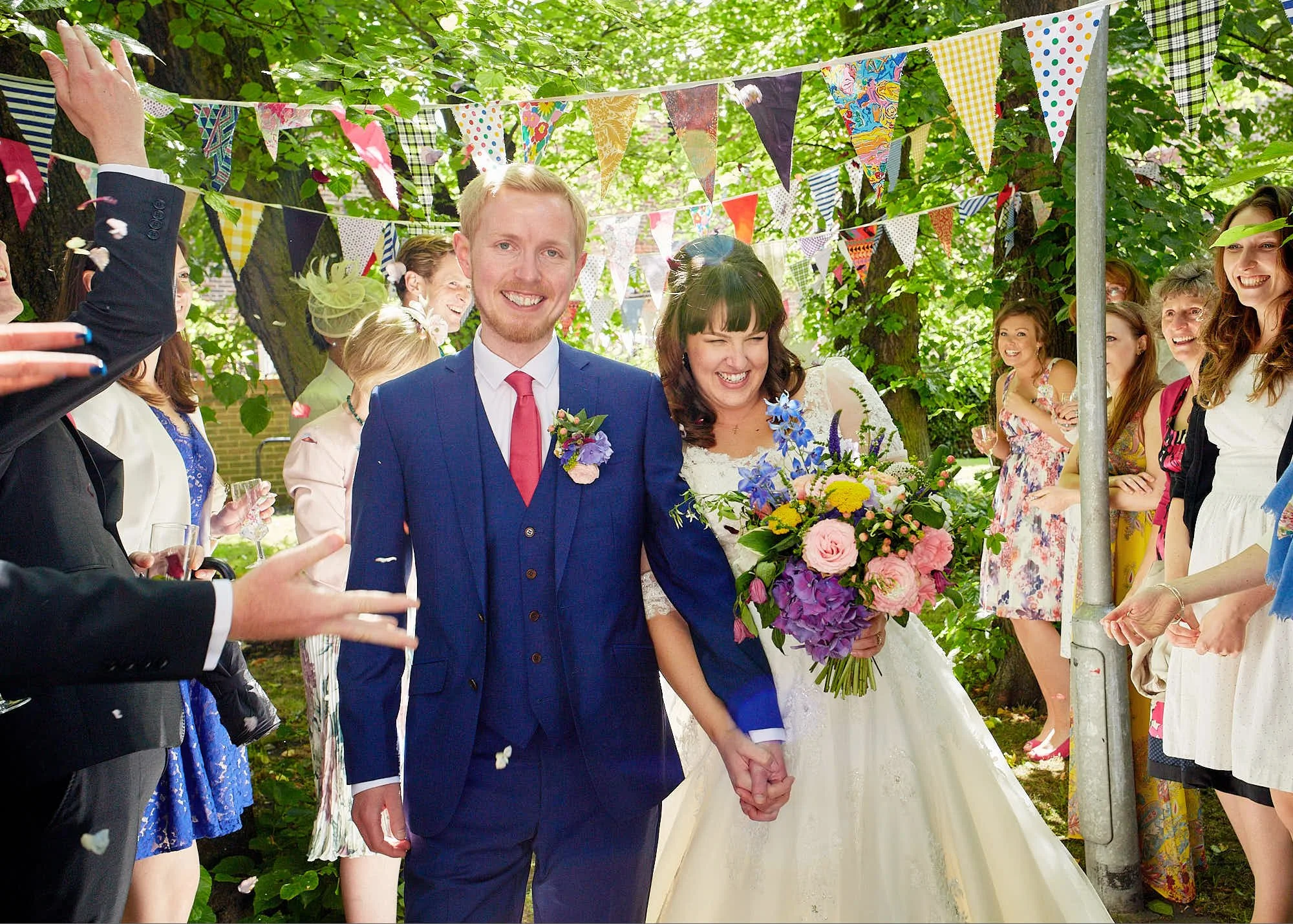 A bride and groom holding hands at an outdoor wedding ceremony, surrounded by smiling guests, with colorful bunting flags hanging from trees, during daytime.
