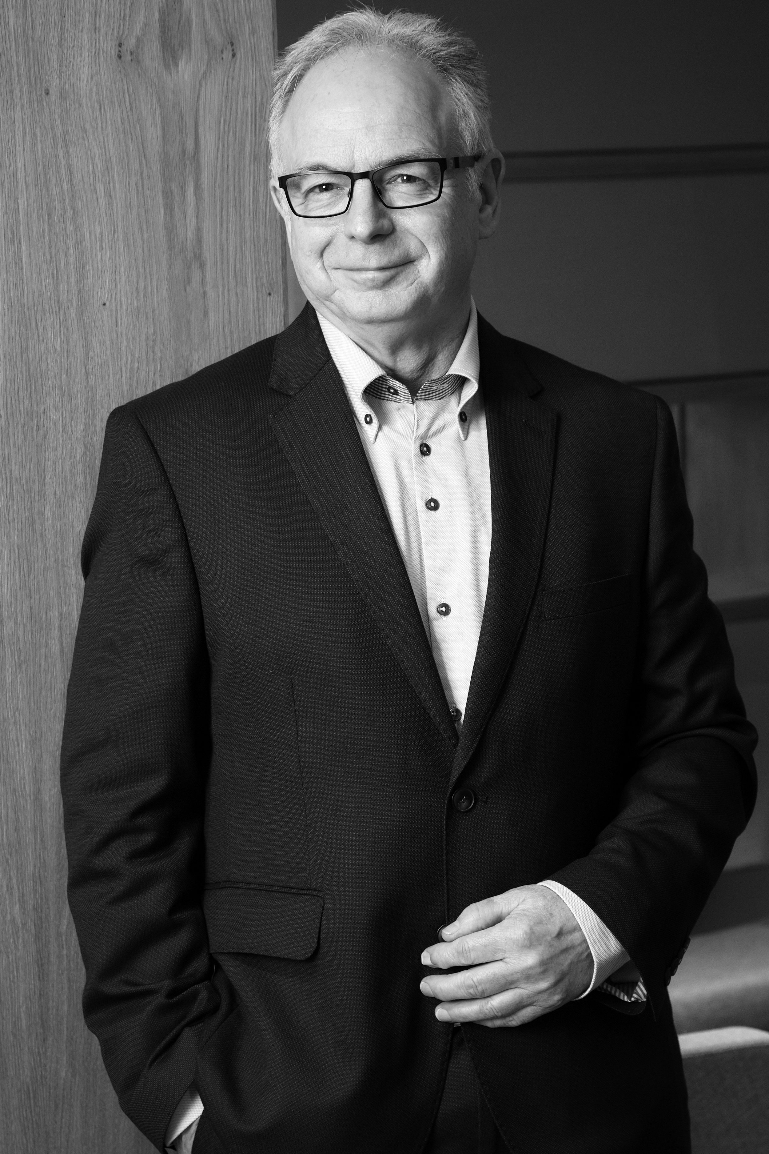A black and white portrait of an older man with glasses, short gray hair, wearing a suit with a button-up shirt, standing next to a wooden wall.