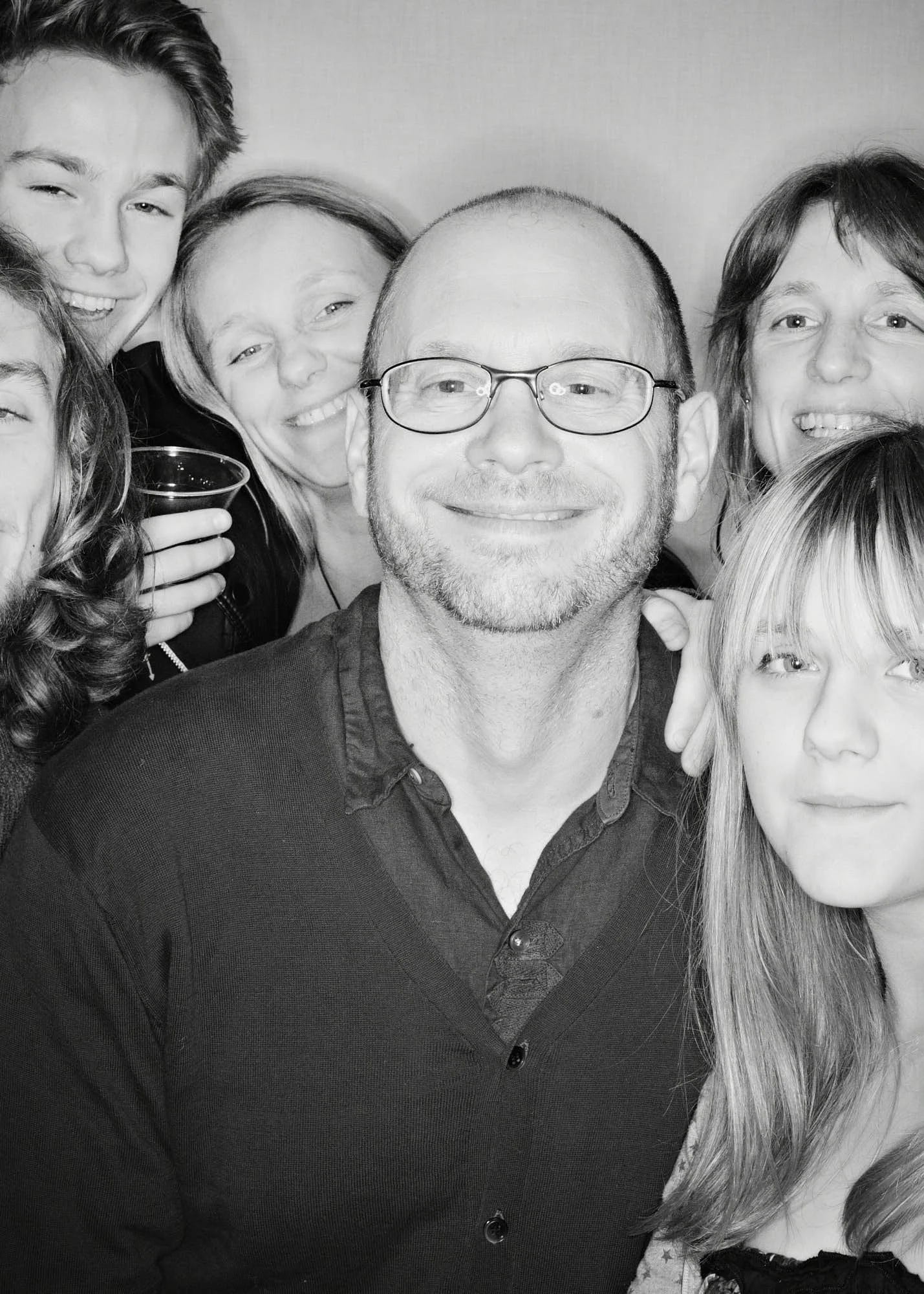 Black and white photo of a group of smiling people, with one man wearing glasses at the center, and some holding drinks.