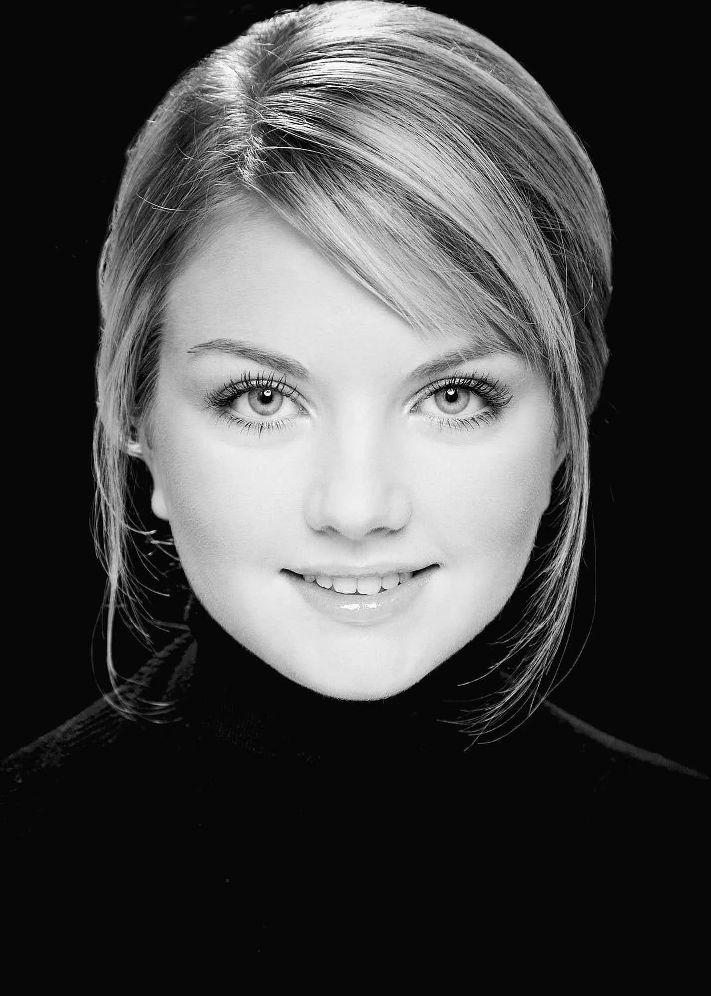 Black and white close-up portrait of a young woman with straight hair, smiling, against a dark background.
