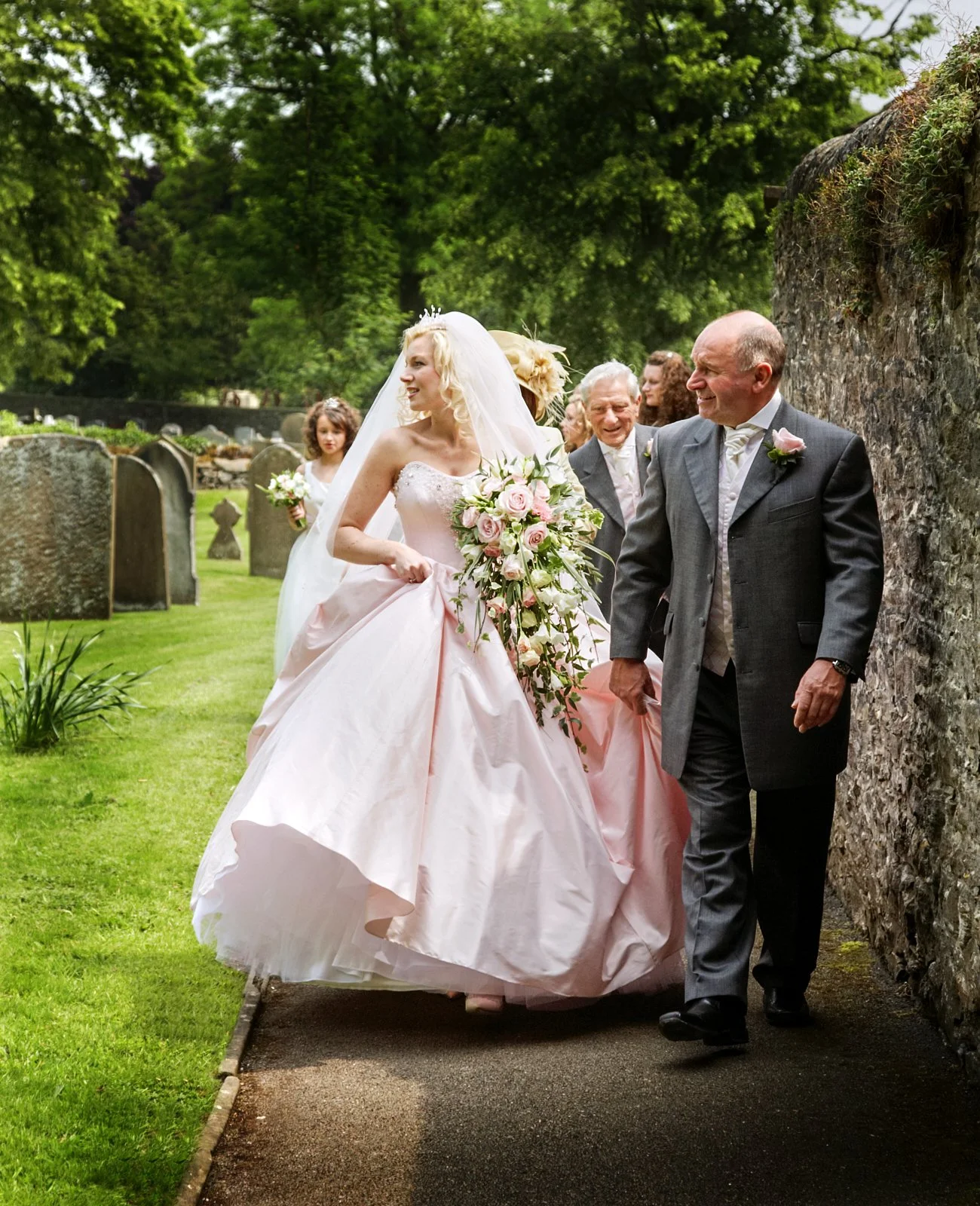 A bride in a pink wedding gown holding a cascading bouquet walking with a groom in a dark suit and light-colored tie in a garden with gravestones, trees, and a stone wall in the background.
