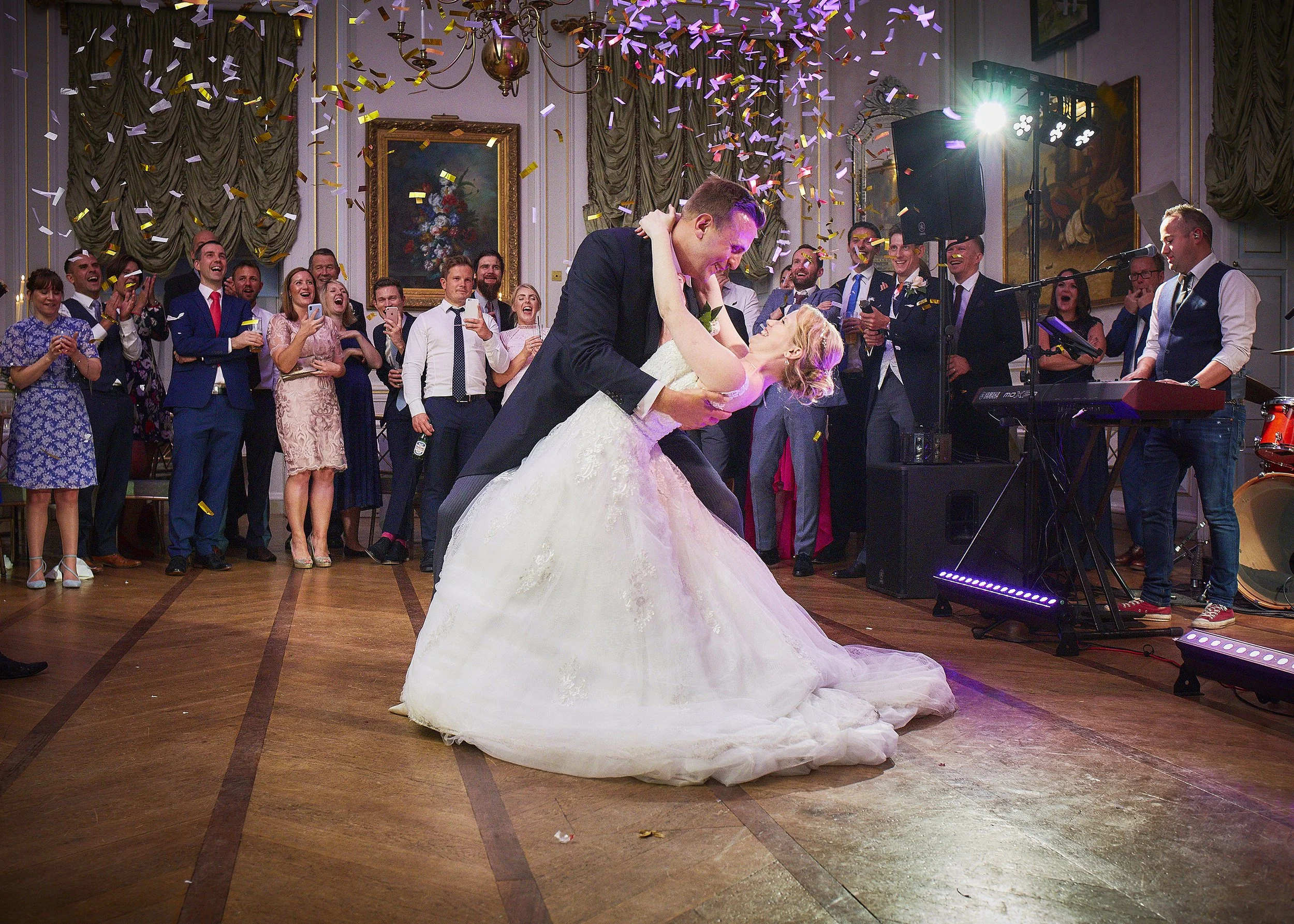A bride and groom are dancing at their wedding reception, surrounded by guests who are smiling and cheering. Colorful confetti is falling from the ceiling, and a candelabra is hanging overhead in an elegant decorated room with paintings on the walls.