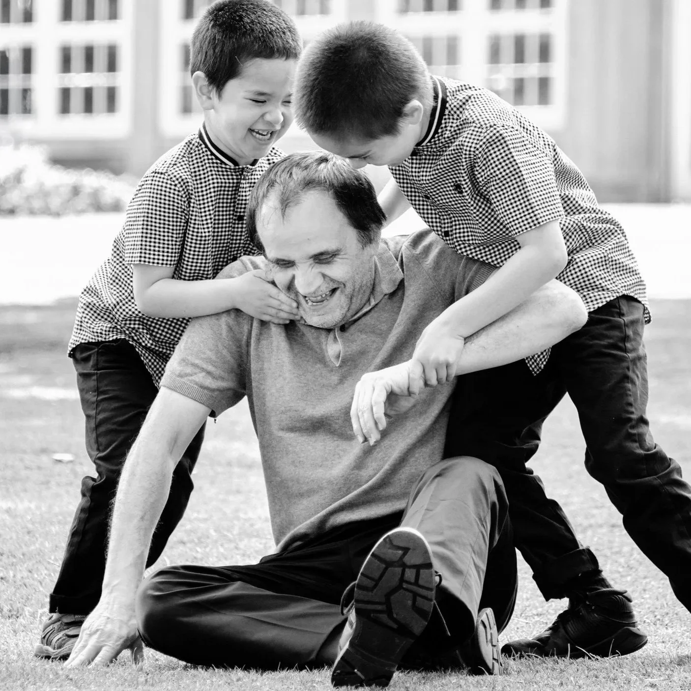 Two young boys and an older man playing and laughing on a grassy field, with the boys pinning the man down in a playful fight, all smiling and enjoying the moment.