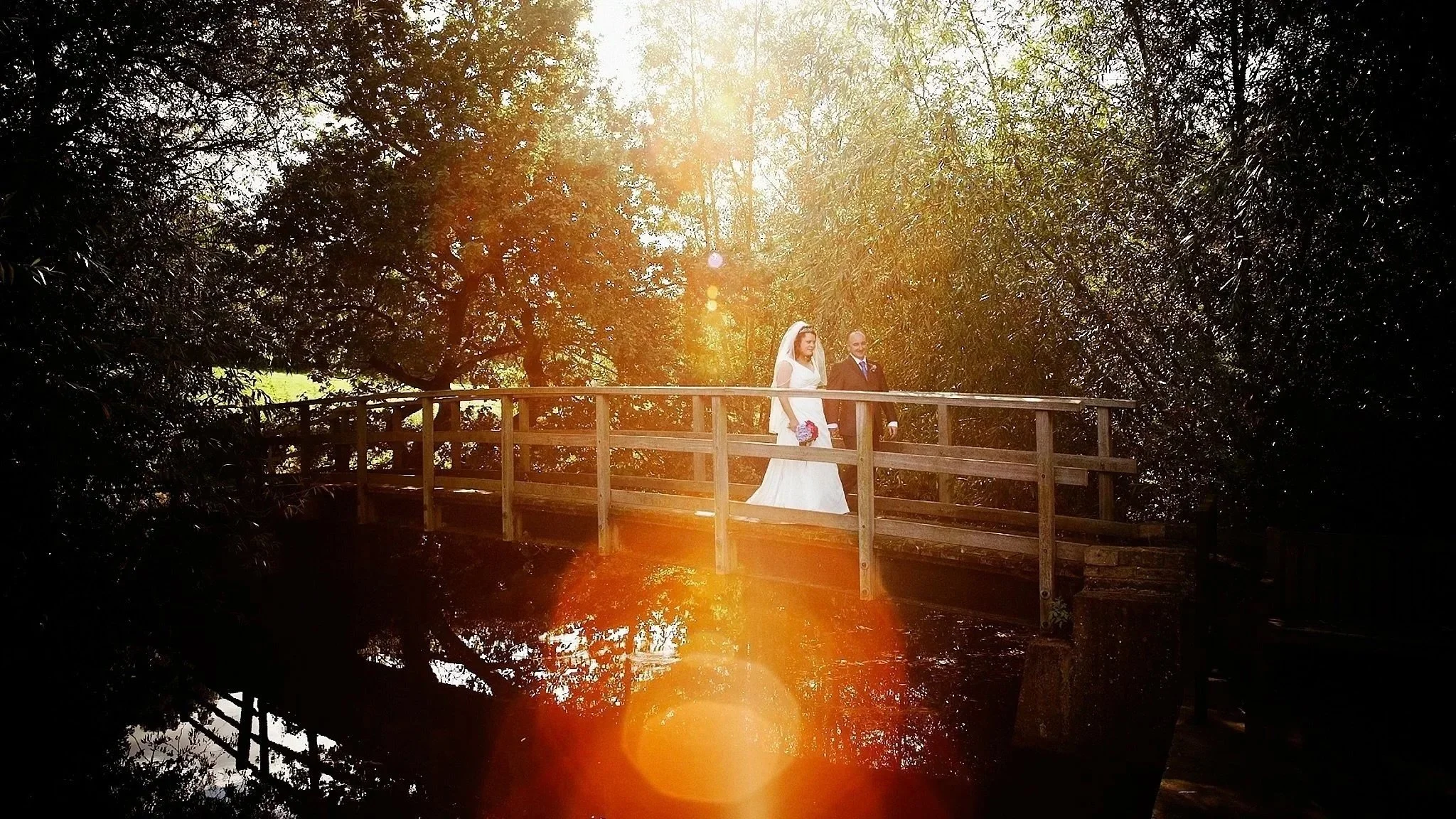 A bride and groom walking on a small wooden bridge in a wooded area with sunlight filtering through the trees.