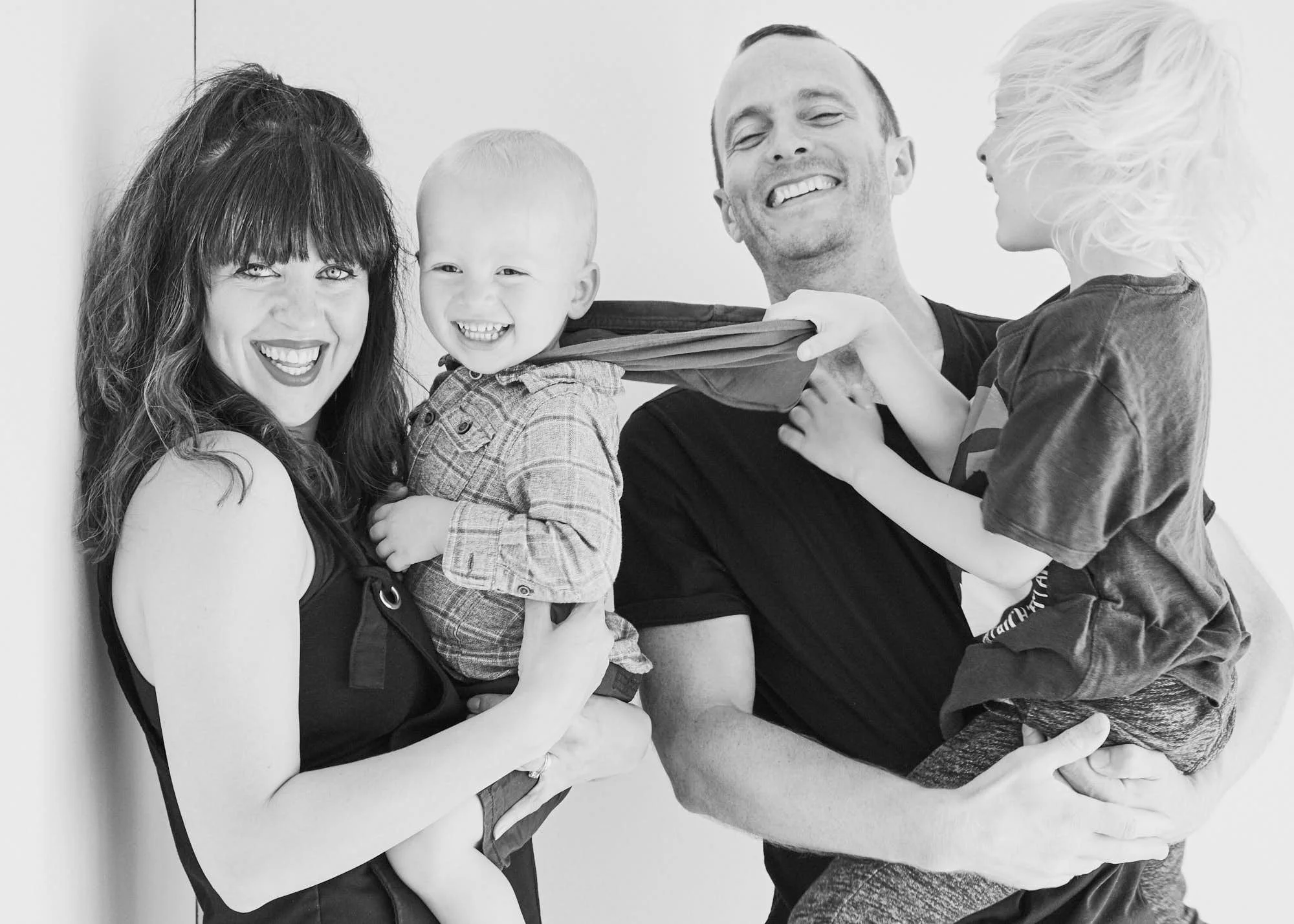 A happy family of four, including a woman, a man, and two children, smiling and playing together indoors in black and white.