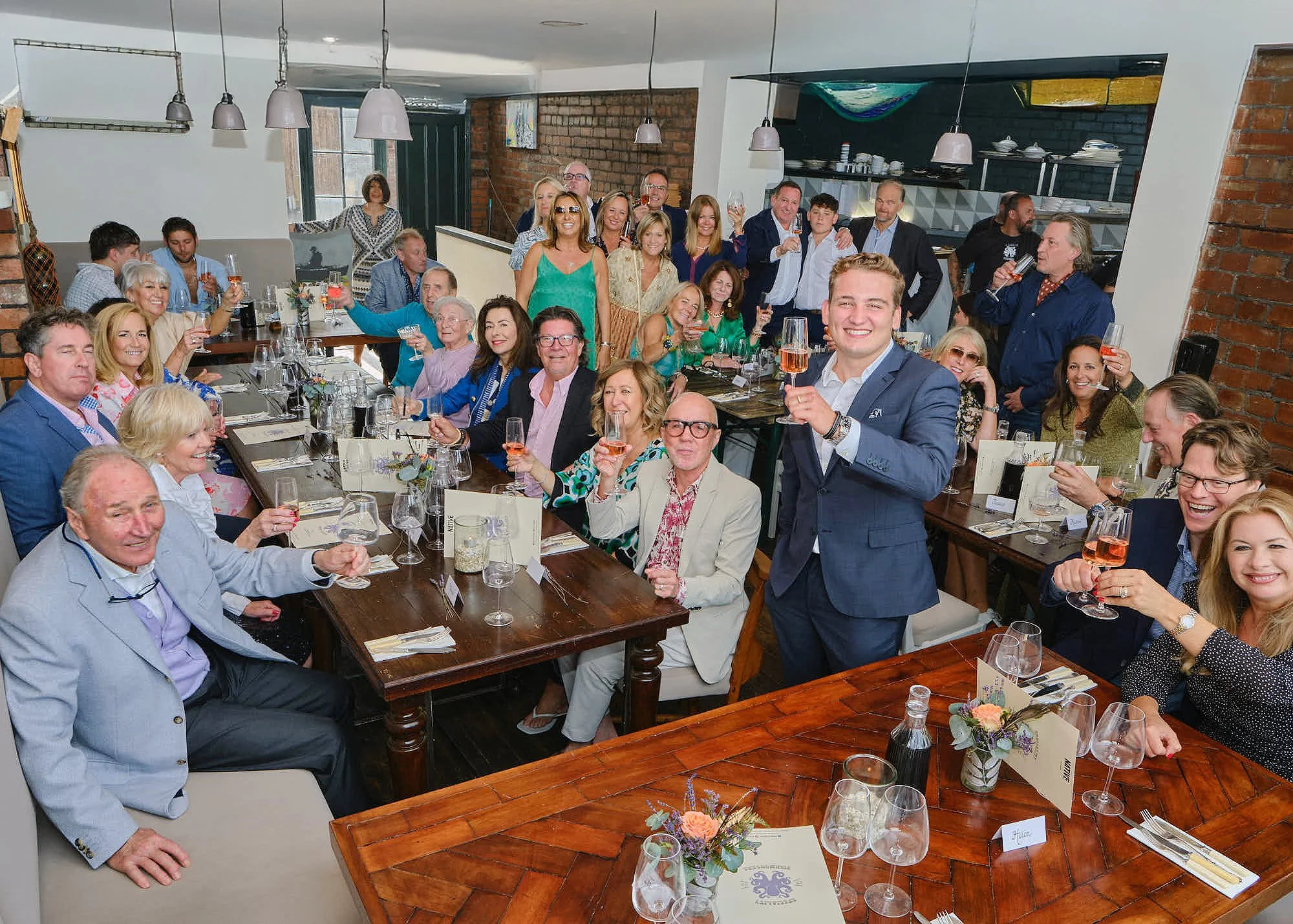 A large group of people gathered in a restaurant, smiling and raising glasses for a toast, with a long table set with wine glasses, flowers, and menus.