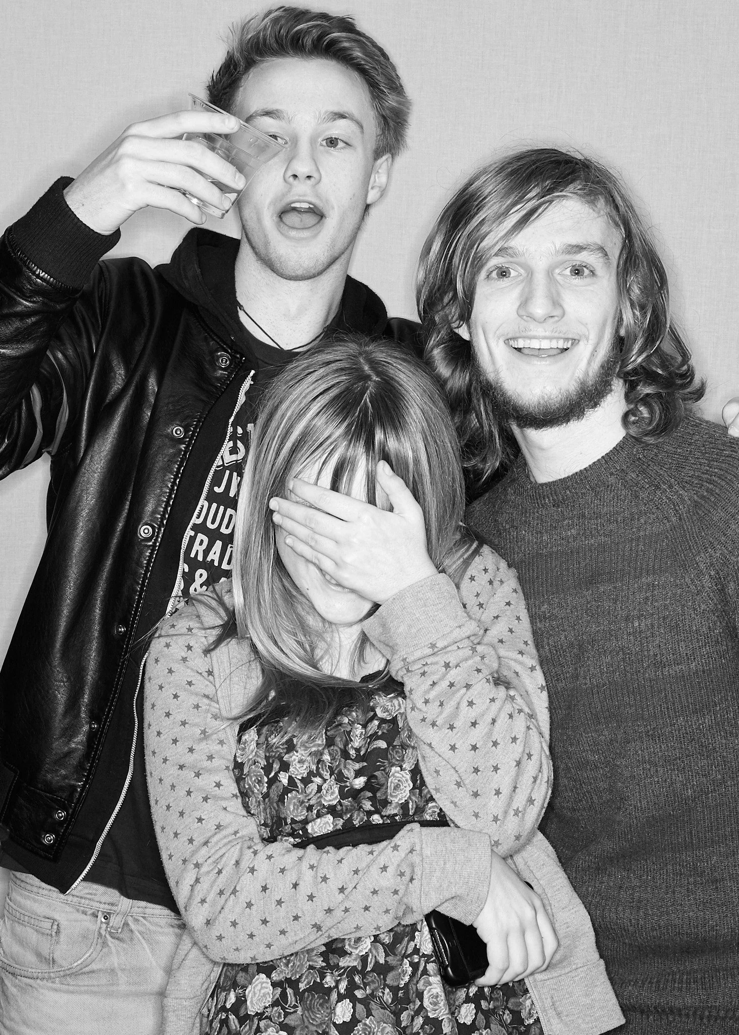 Three young people in a photo, one covering her face, one holding a drink, and one smiling. The girl is wearing a floral dress and the guys are dressed casually.