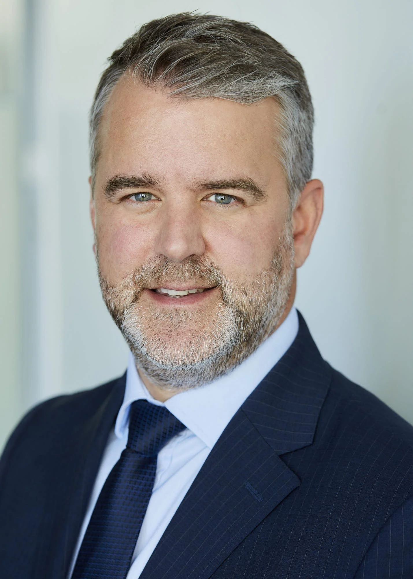 Close-up of a middle-aged man with gray hair and a beard, wearing a dark suit, white shirt, and blue tie, smiling slightly.