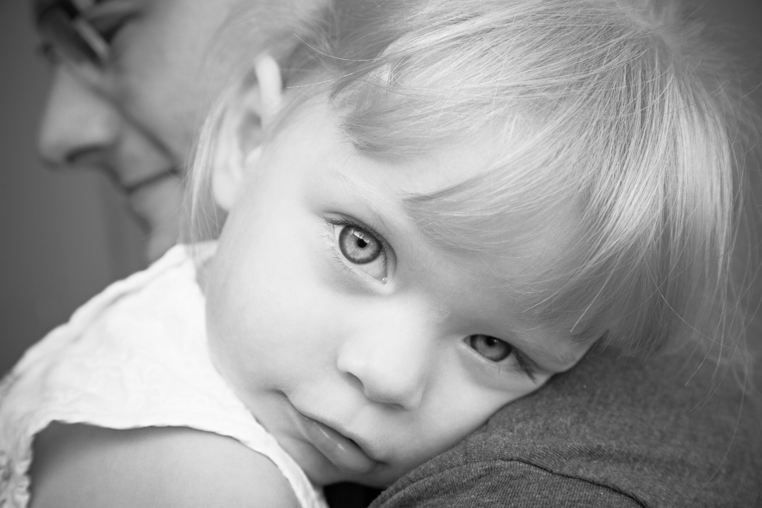 A black and white close-up photograph of a young child with light-colored hair and bright eyes, lying down and looking directly at the camera.
