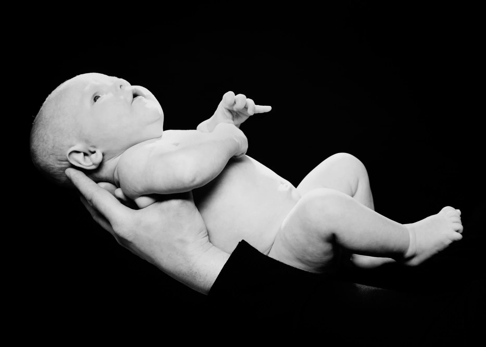 A black-and-white photo of a newborn baby being held gently in an adult's hands against a black background.