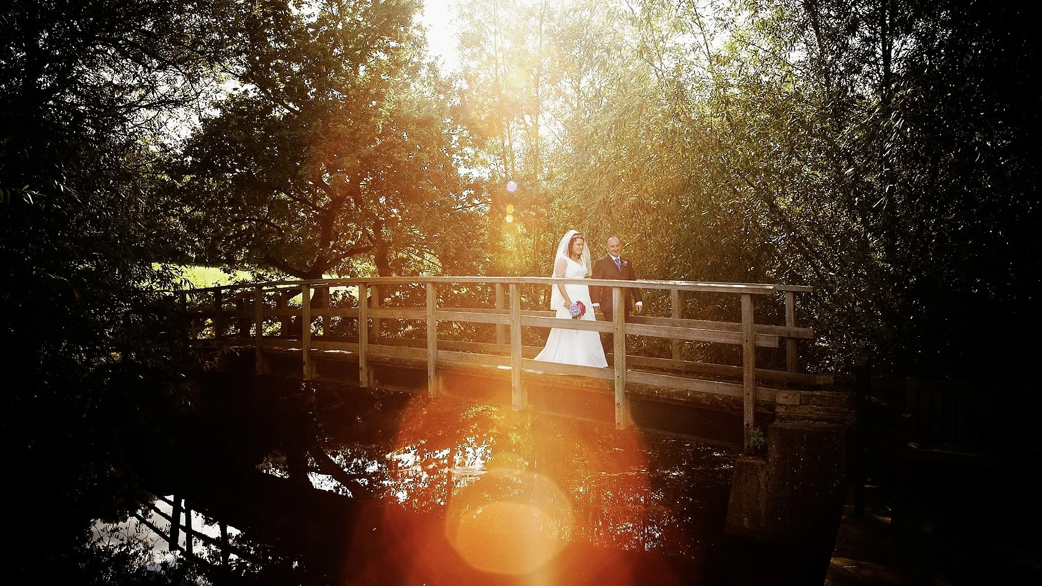 A bride and groom walking on a wooden bridge surrounded by trees, with sunlight shining through the foliage.
