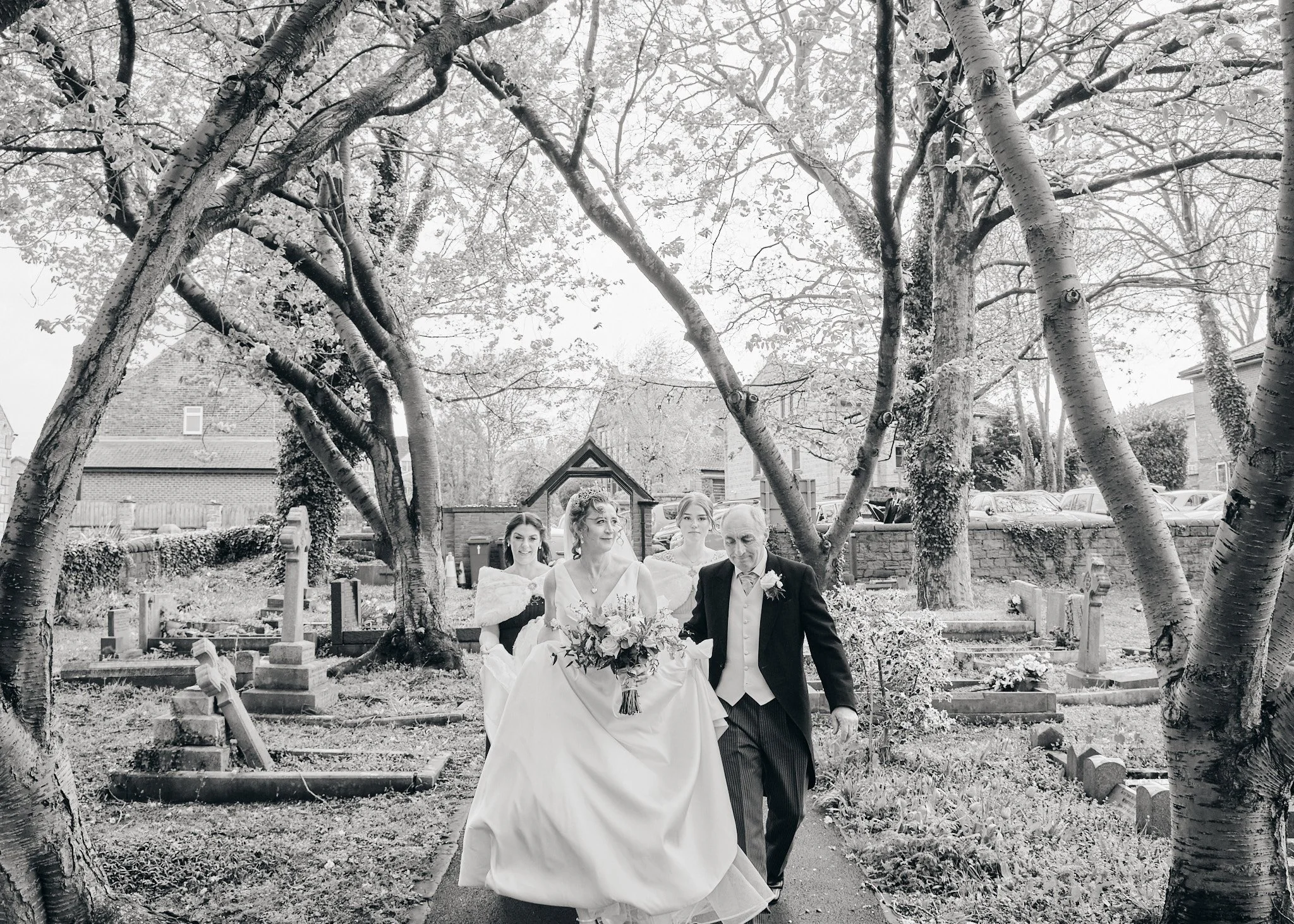 A wedding procession walking through a garden with trees, with a bride in a white gown holding a bouquet, and an older man in a suit.