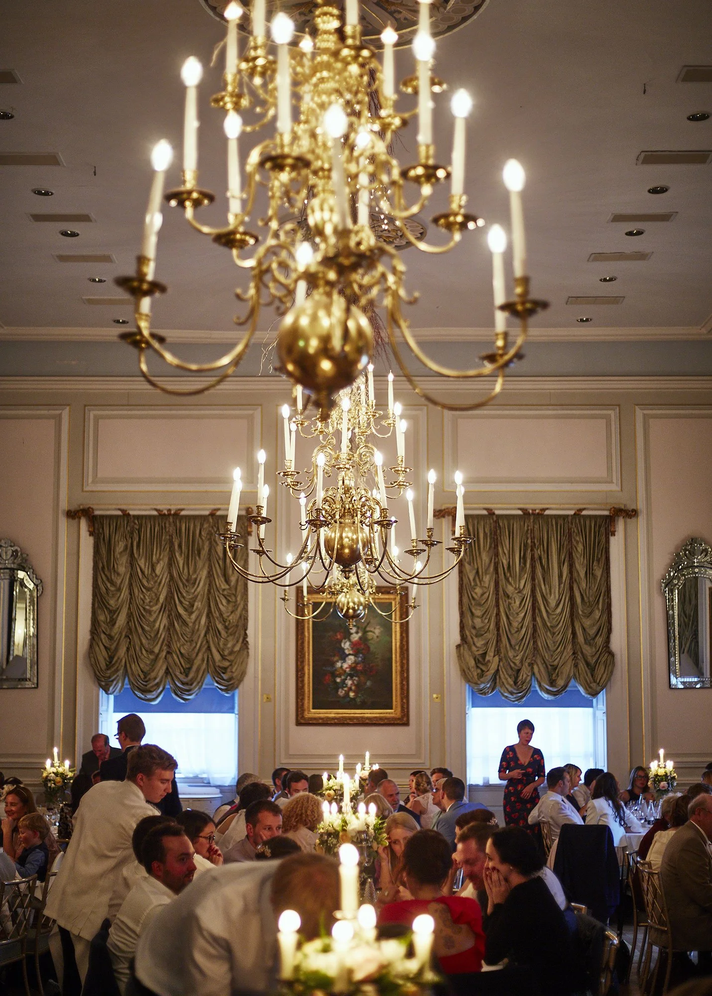 Elegant dining room filled with people seated at tables, illuminated by multiple grand gold chandeliers hanging from the ceiling, with tall windows draped with curtains and framed artwork on the walls.