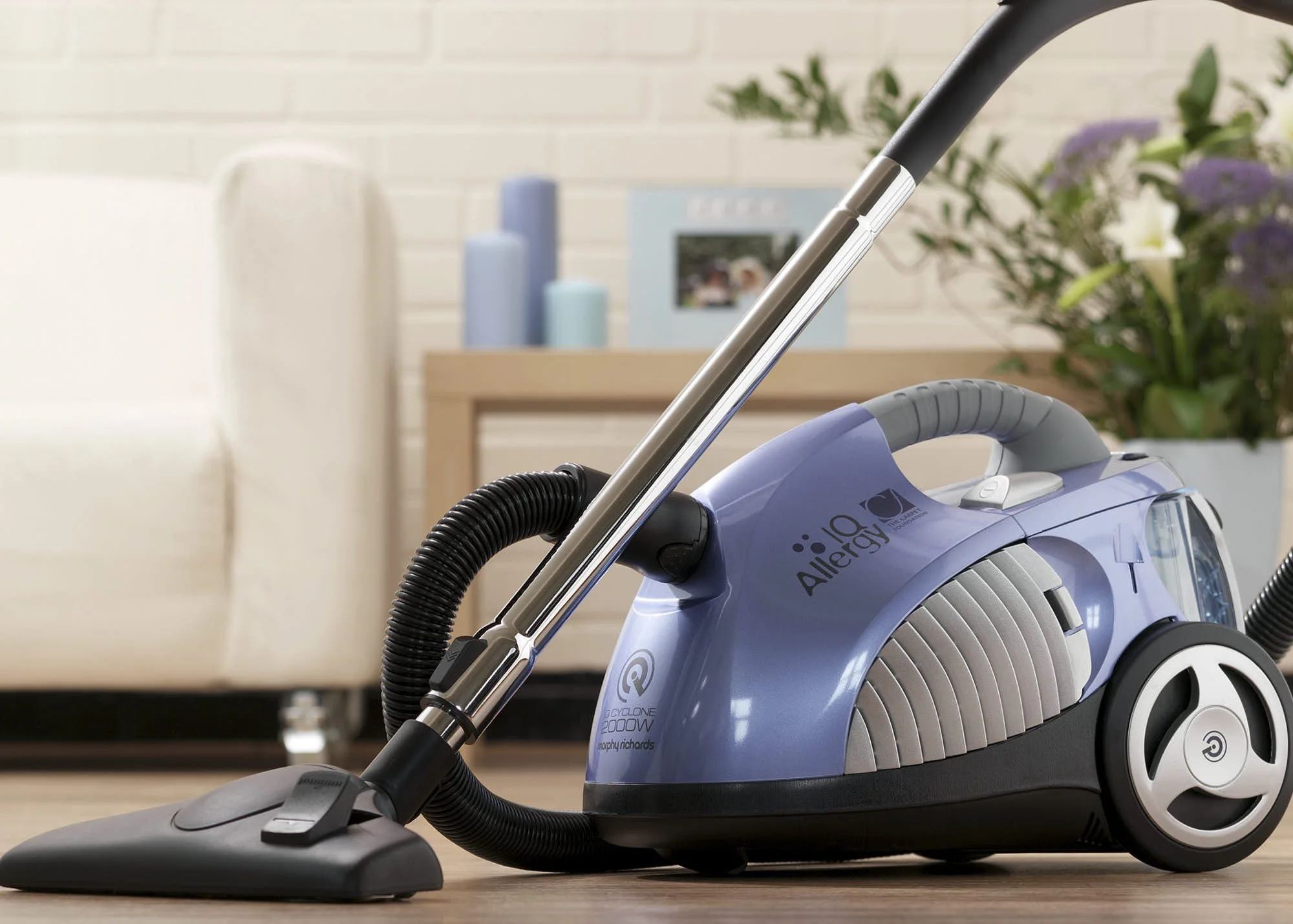 A blue industrial vacuum cleaner with a black hose and a vacuum head, placed on a wood floor, with a background of a light-colored wall, a bookshelf with blue and purple items, and a bouquet of purple and yellow flowers.