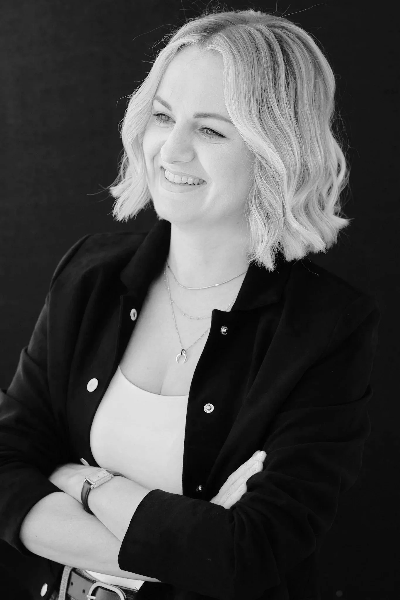 Black and white portrait of a woman with shoulder-length wavy blonde hair, smiling and looking to her right. She is wearing a dark jacket over a light top, with her arms crossed and a watch on her left wrist.
