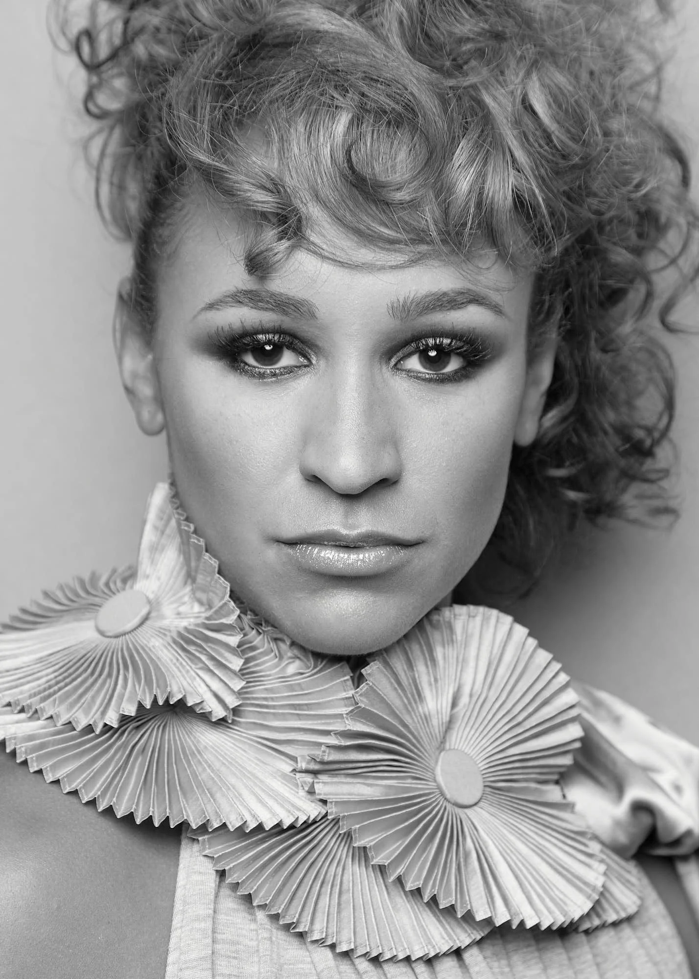 Black and white close-up portrait of a woman with curly hair, wearing earrings and a ruffled collar, looking directly at the camera.