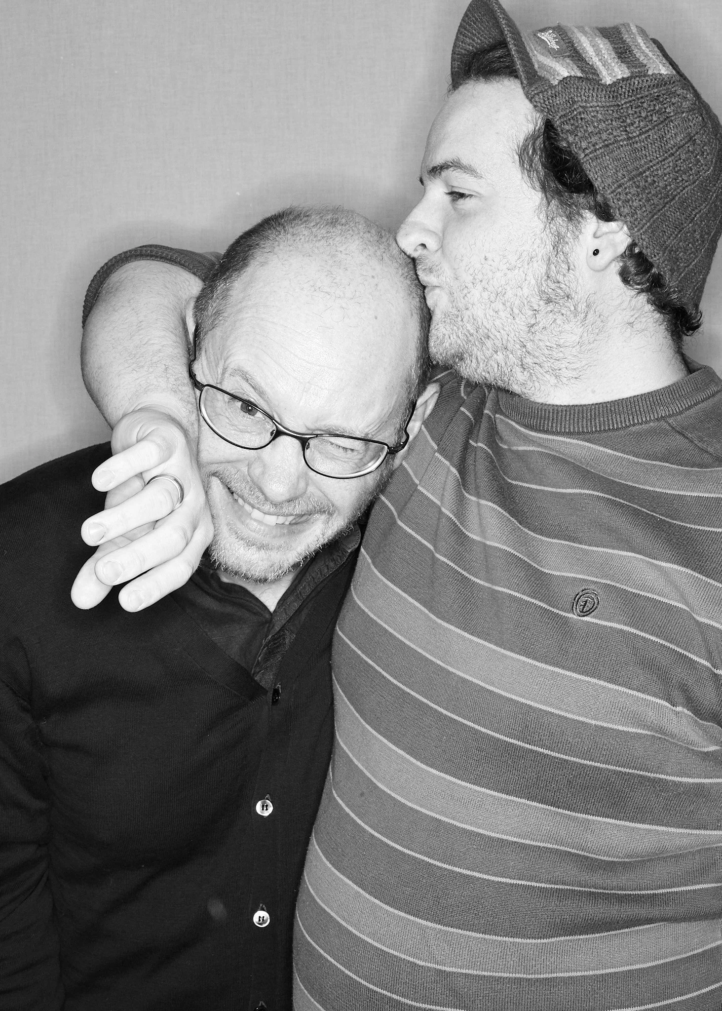 Two men hugging and sharing a kiss on the forehead. One man has glasses and is smiling, the other is wearing a striped shirt and a beanie.