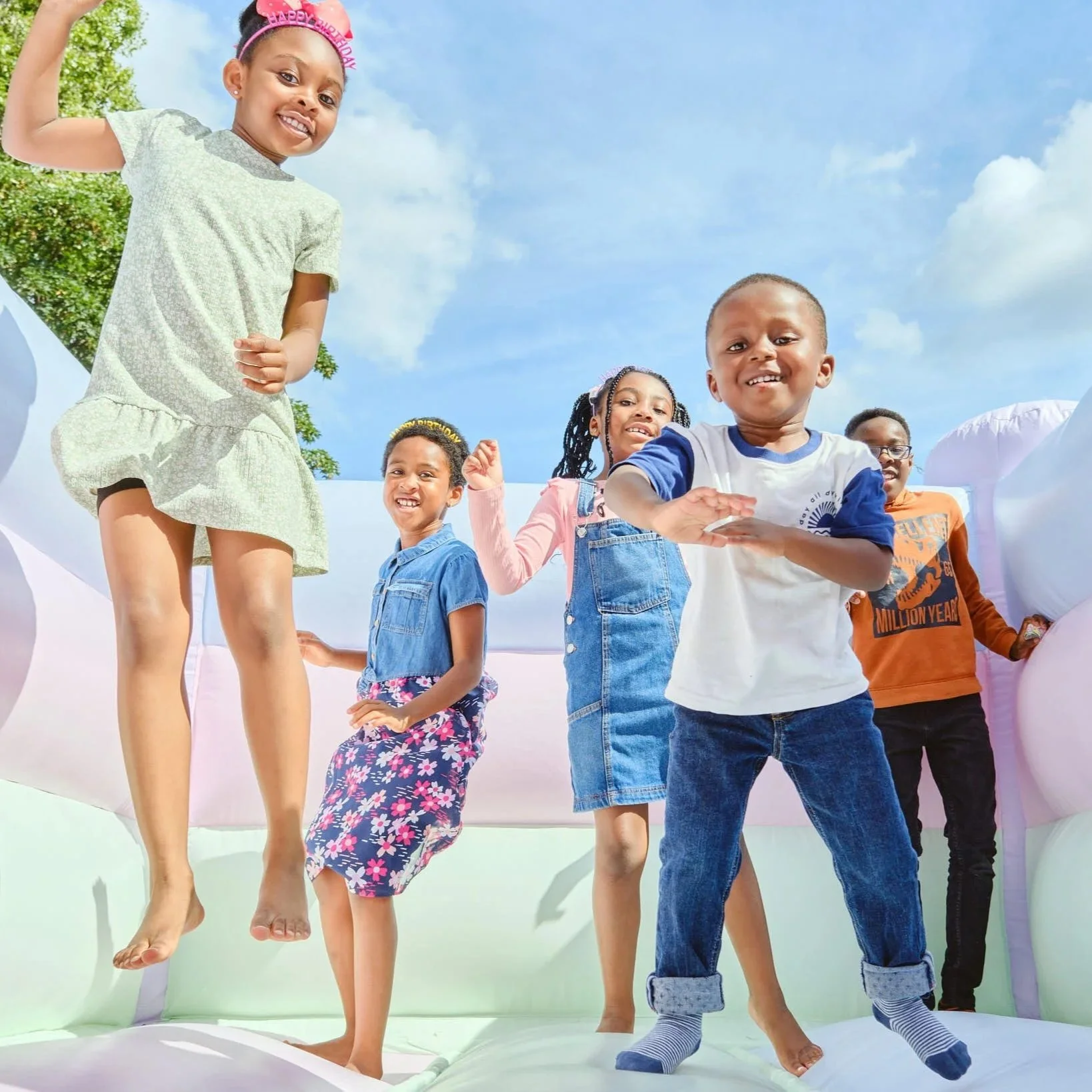 Group of children celebrating a birthday on an inflatable bounce house, with a blue sky and clouds in the background.