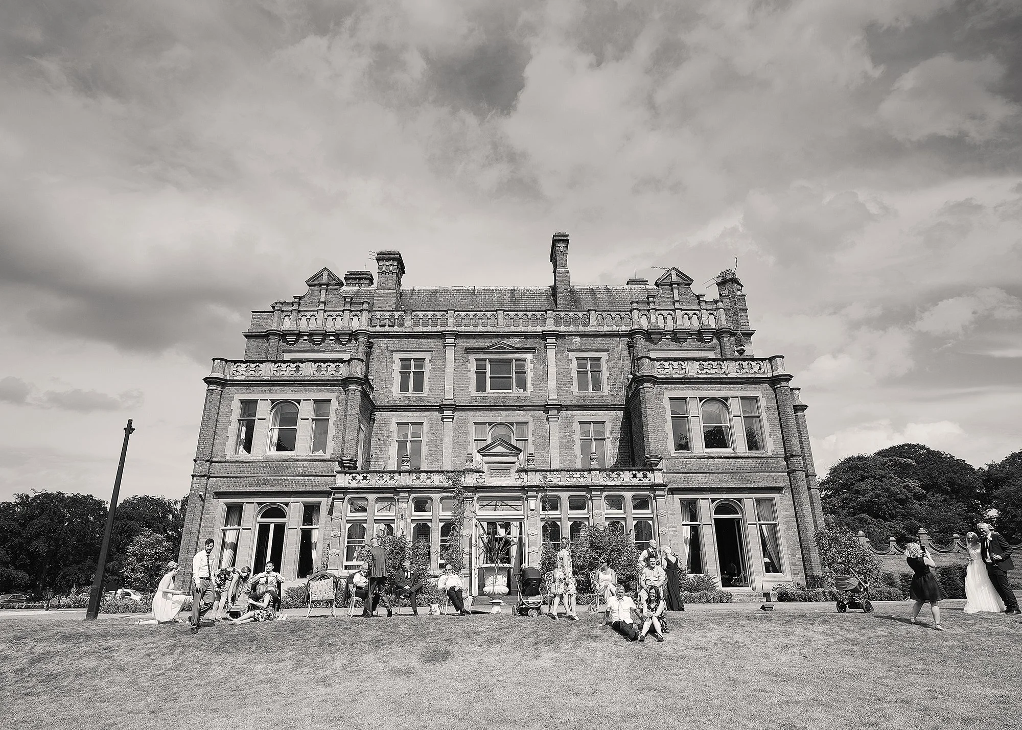 A large, ornate stone building with multiple windows and decorative architectural details. In front, there are many people gathered on the lawn, some sitting, walking, and standing, possibly at an event or gathering. The sky above is cloudy.
