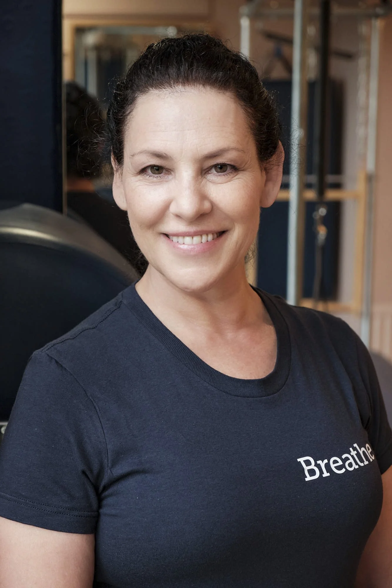 A woman with dark curly hair tied back, smiling, wearing a navy blue t-shirt with the word 'Breathe' on it, in an indoor setting with shelves and exercise equipment in the background.