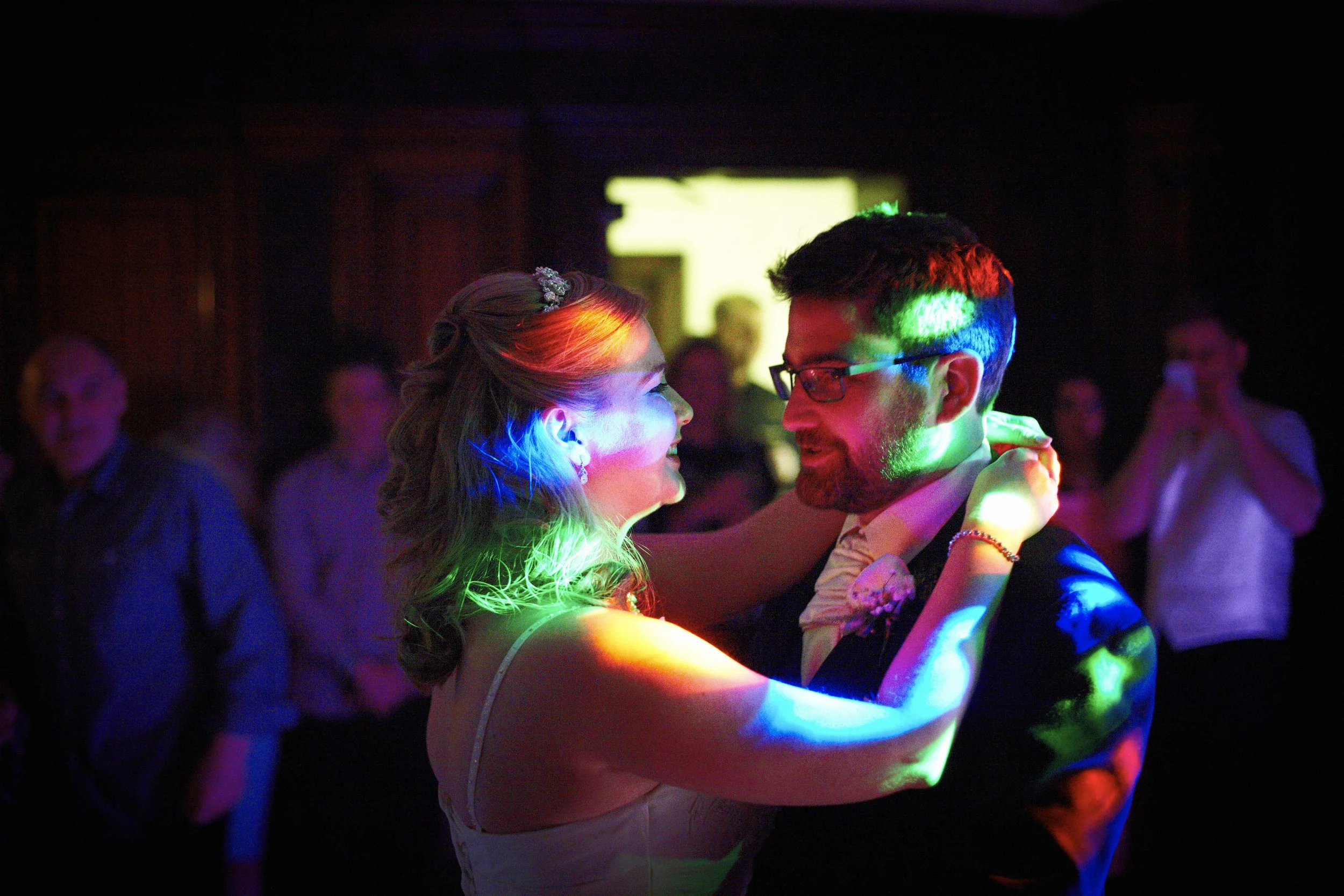 A couple dancing closely at a dimly lit party, illuminated by colorful stage lights, with other guests in the background.