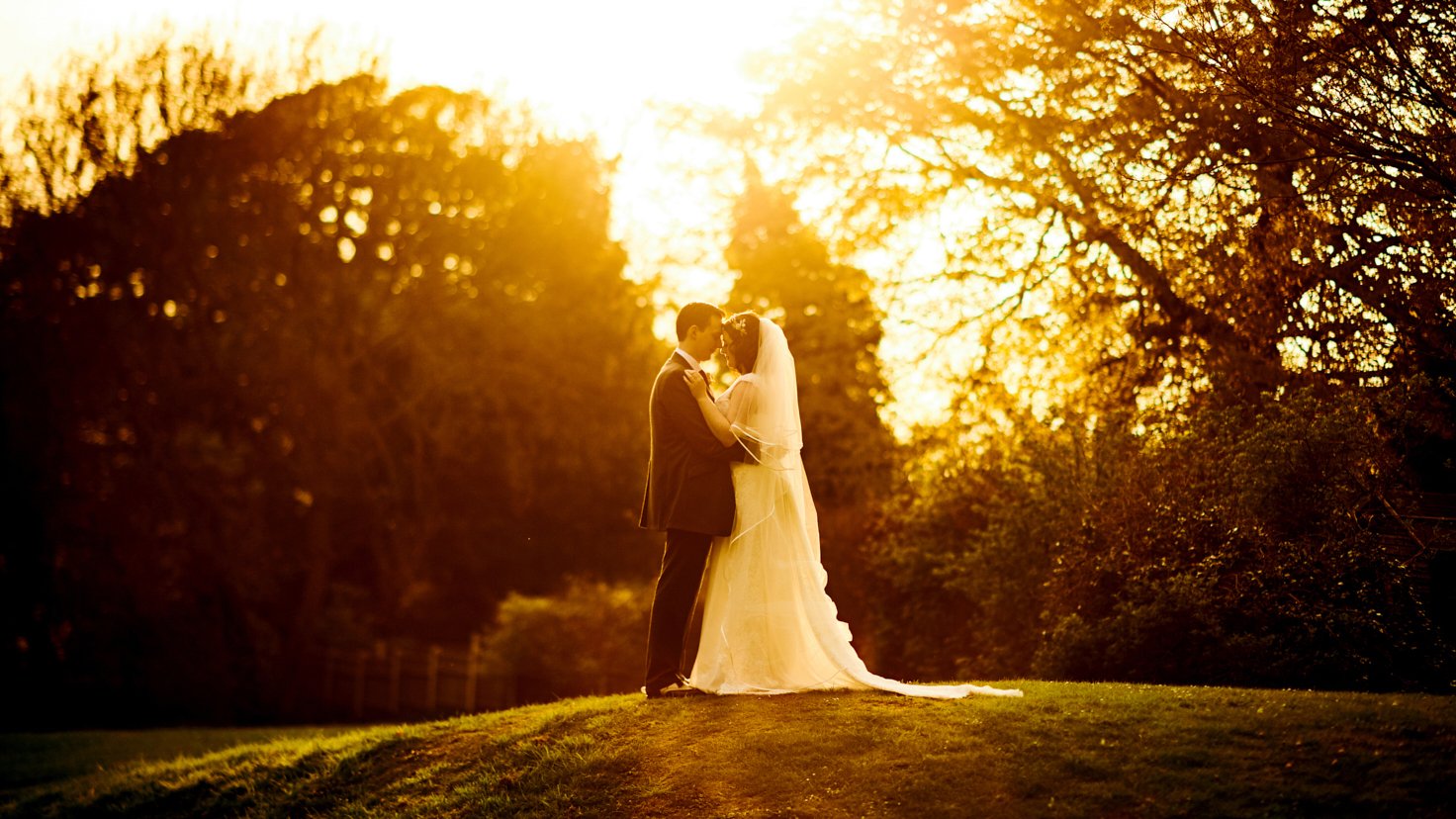 A bride and groom in wedding attire standing close together on a grassy hill, embracing during sunset with backlit trees in the background.