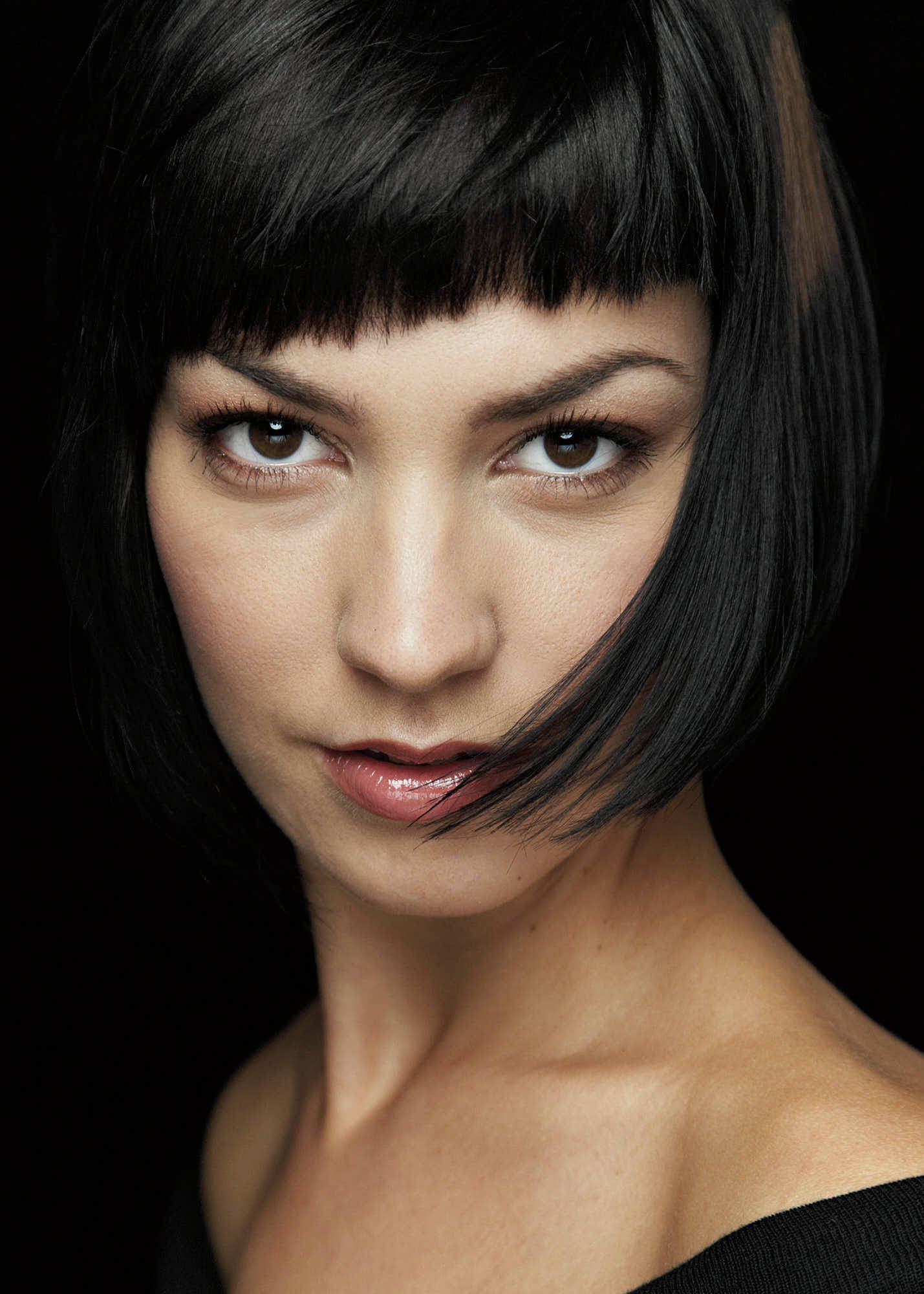 Close-up portrait of a woman with black hair, styled in a bob with bangs, and brown eyes, wearing makeup with natural tones, against a dark background.