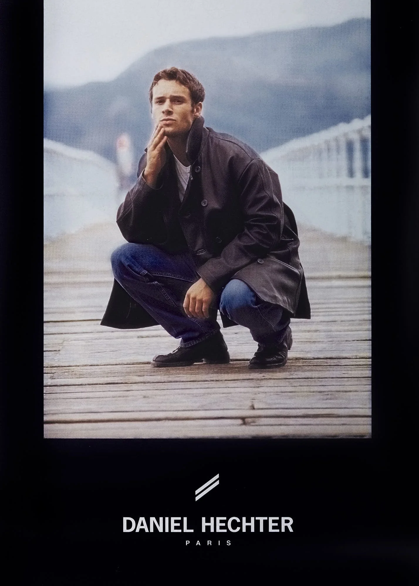 A young man in a black leather jacket and jeans crouching on a wooden pier with a bridge and mountains in the background.