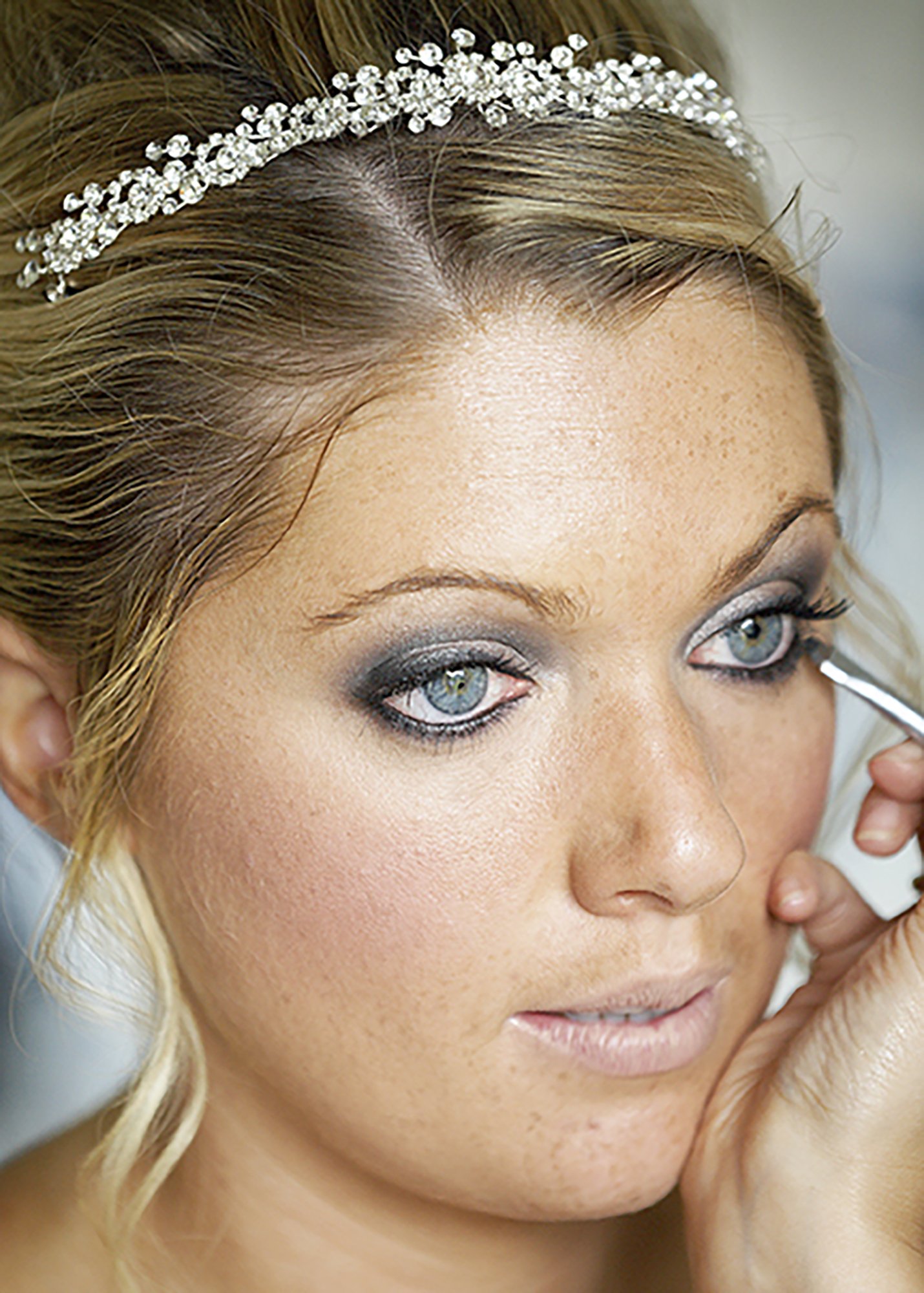 Close-up of a woman with blonde hair, blue eyes, makeup, and a rhinestone headband, touching her face gently.