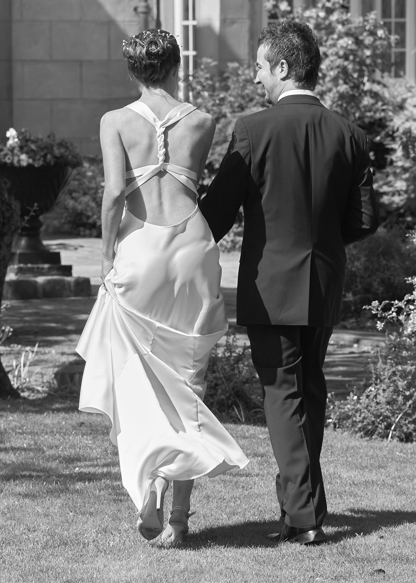 A bride and groom dancing outdoors on a lawn, with the bride in a backless wedding dress and the groom in a suit, surrounded by greenery and a stone building.