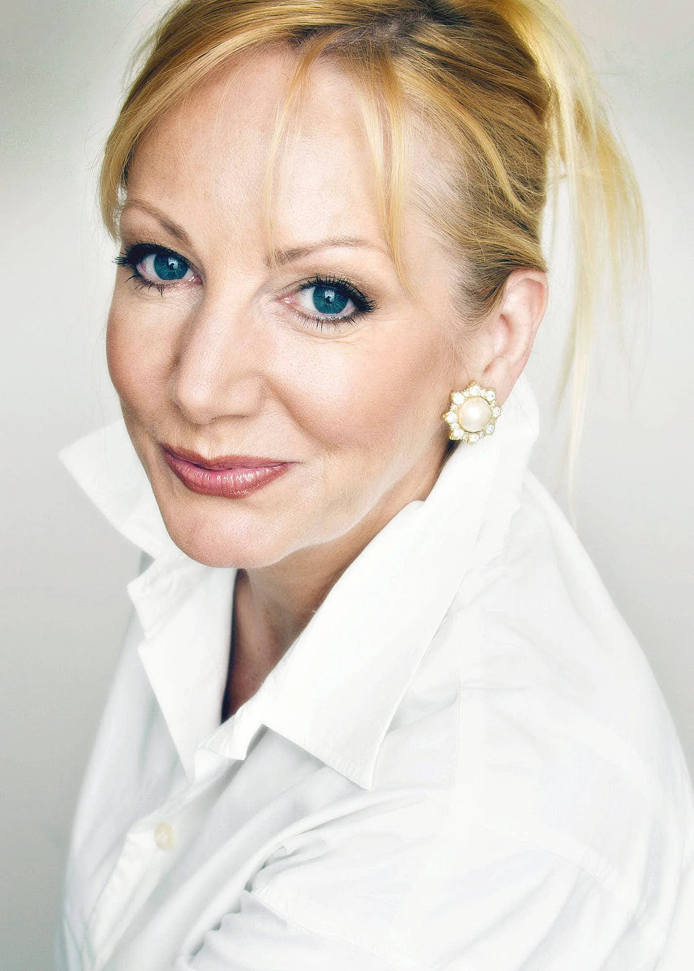 A woman with blonde hair styled in a loose updo, bright blue eyes, light makeup, pearl earrings with gold details, and wearing a white shirt, smiling softly.