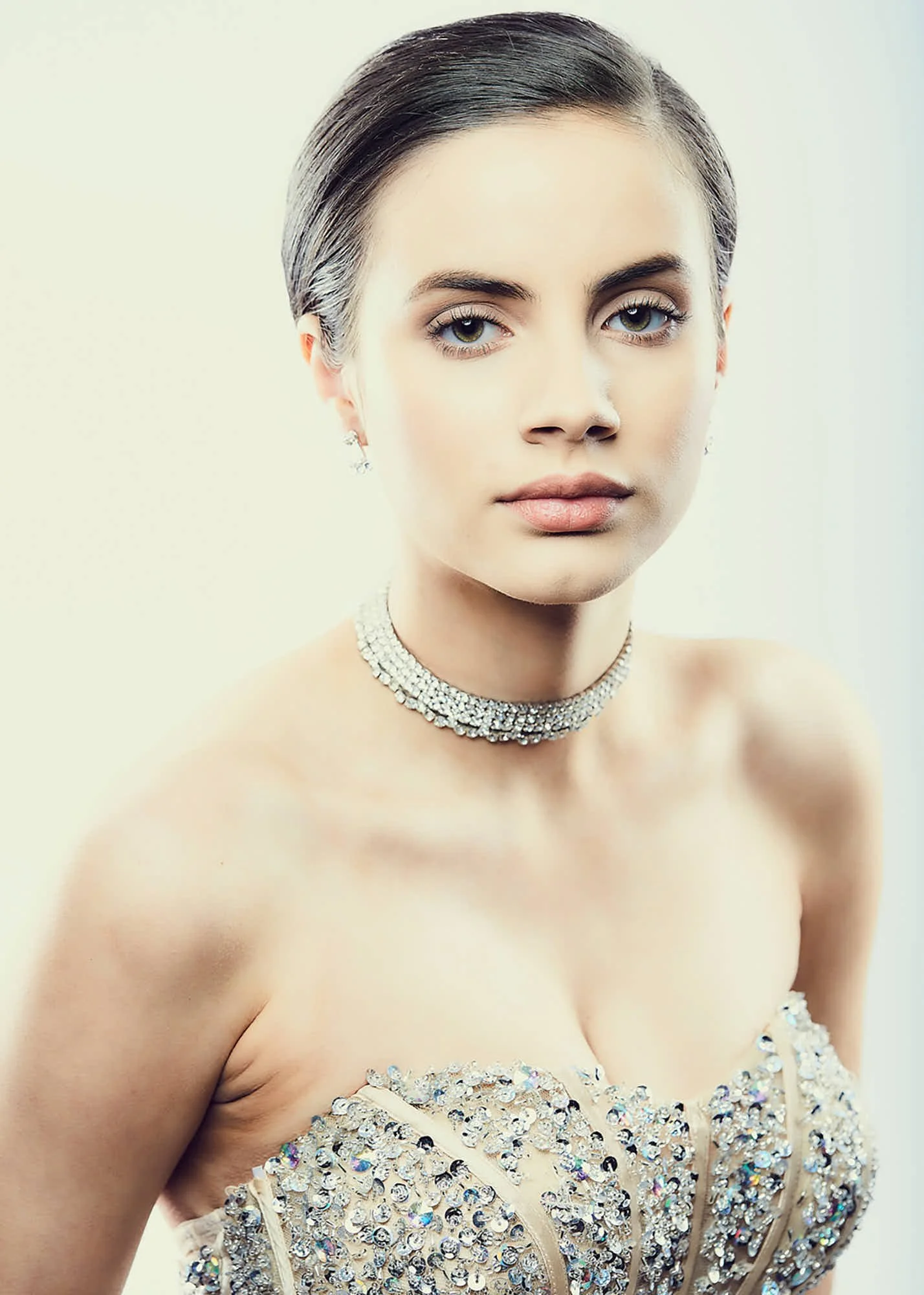 Close-up of a woman with short dark hair, wearing diamond jewelry and a dress adorned with sequins, with a neutral background.