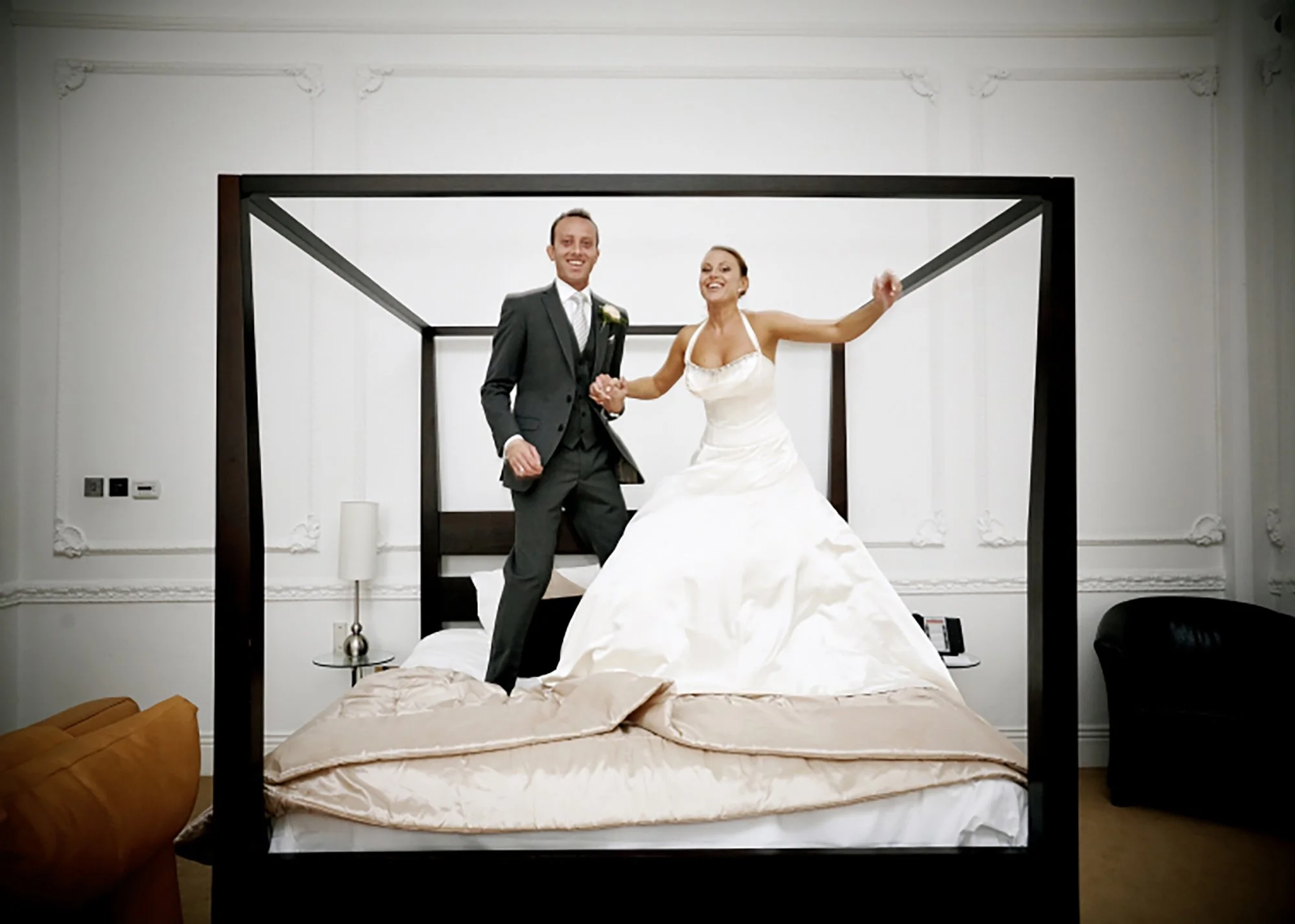 A newlywed couple standing on a bed, holding hands and smiling, inside a modern, minimalist room with white walls and decorative crown molding.