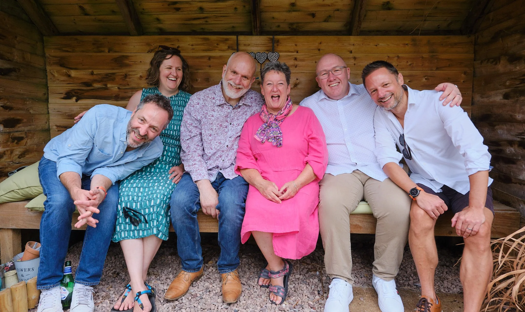 A group of seven adults, four men and three women, sitting and leaning on a wooden bench outdoors. They are smiling and laughing, dressed casually, in front of a wooden wall with decorative hearts hanging.