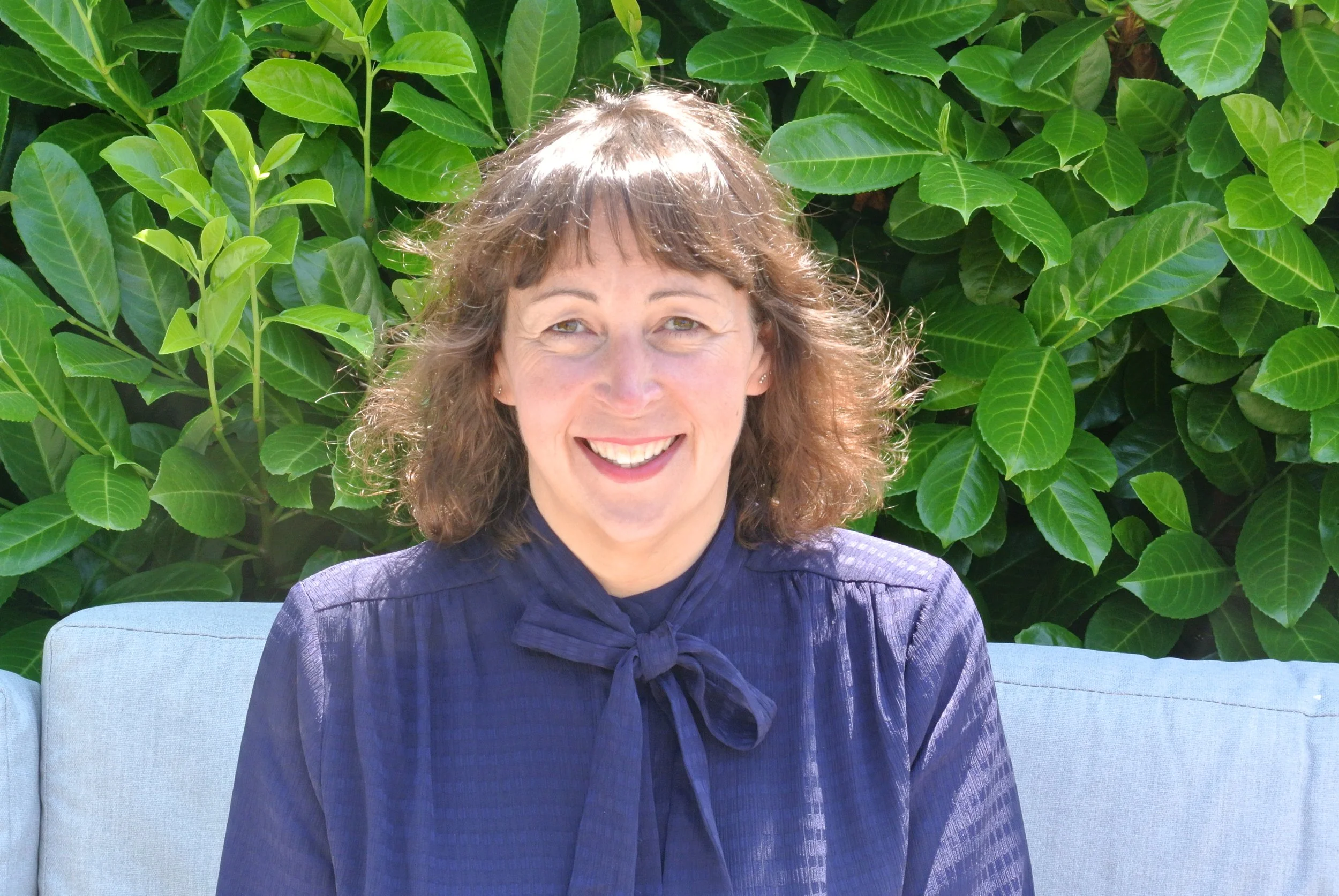 A woman with curly brown hair is smiling and sitting on a light blue couch. She is wearing a dark purple blouse with a tied bow at the collar. Behind her is a large green leafy plant.