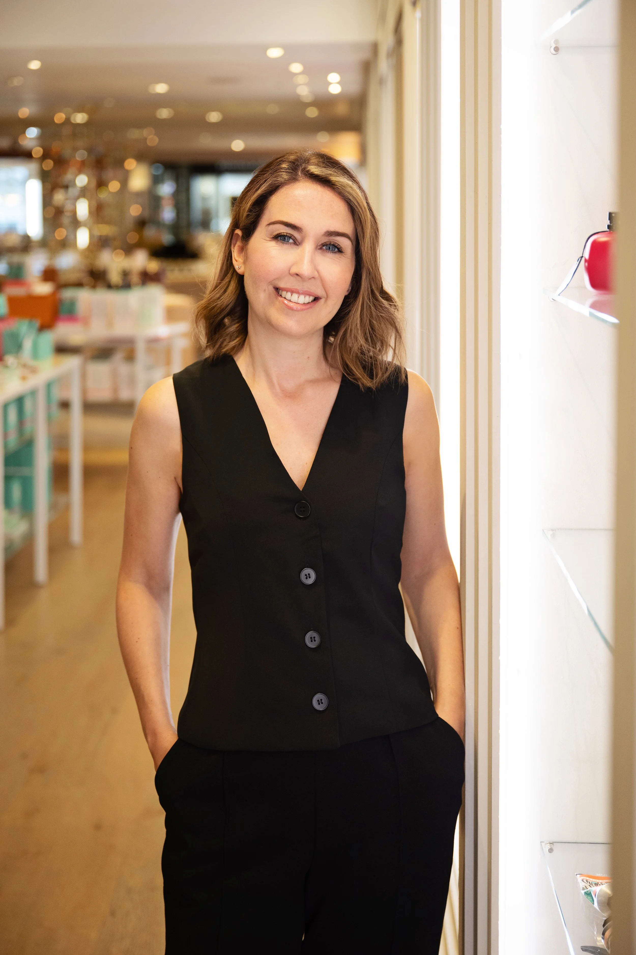 A woman with shoulder-length brown hair and blue eyes, smiling, wearing a sleeveless black button-up top, standing in a well-lit room with shelves and tables in the background.