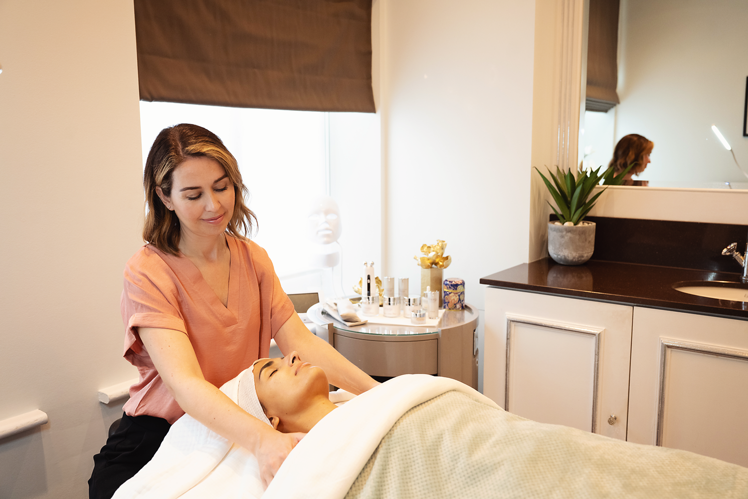 A woman receives a facial treatment from an esthetician in a spa-like setting with skincare products and a potted plant on the counter.